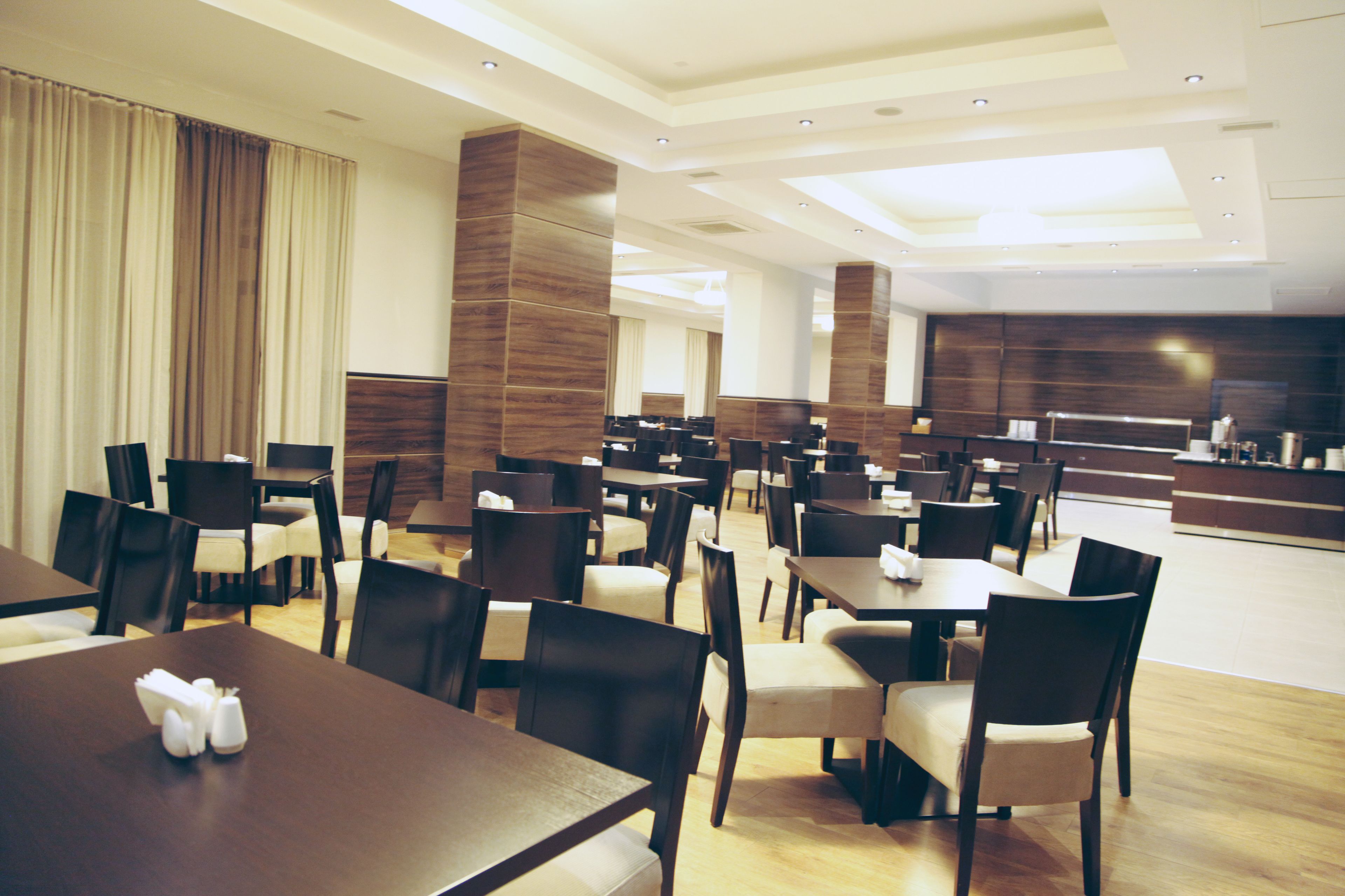 Un moderno hall de hotel con varias mesas y sillas para comer, una zona de recepción y un amplio ventanal con cortinas.