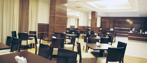 A modern hotel lobby with multiple dining tables and chairs, a reception area, and a large window with curtains.
