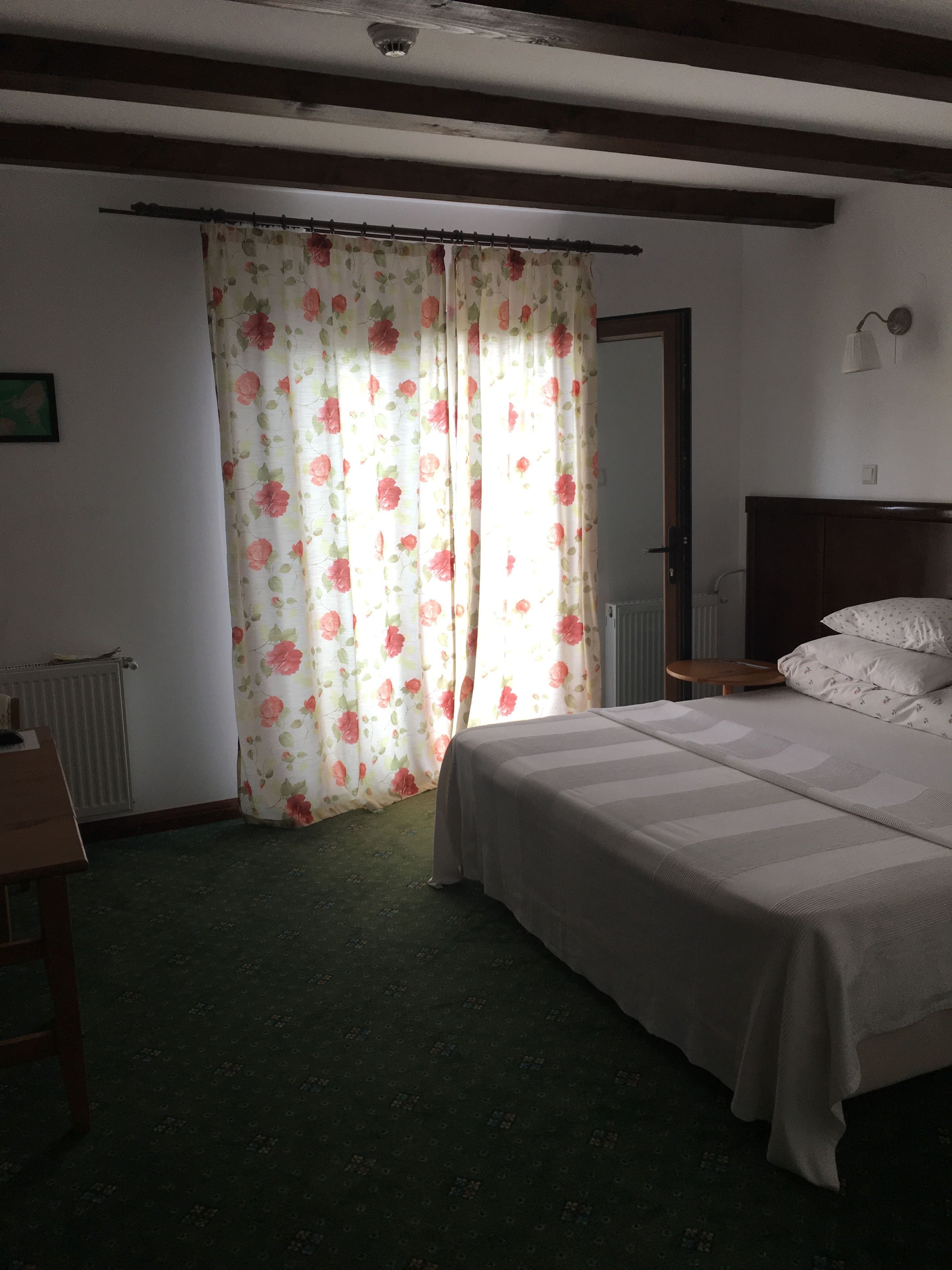 A bedroom with a bed, a wooden headboard, a nightstand, a lamp, and a window with floral curtains.