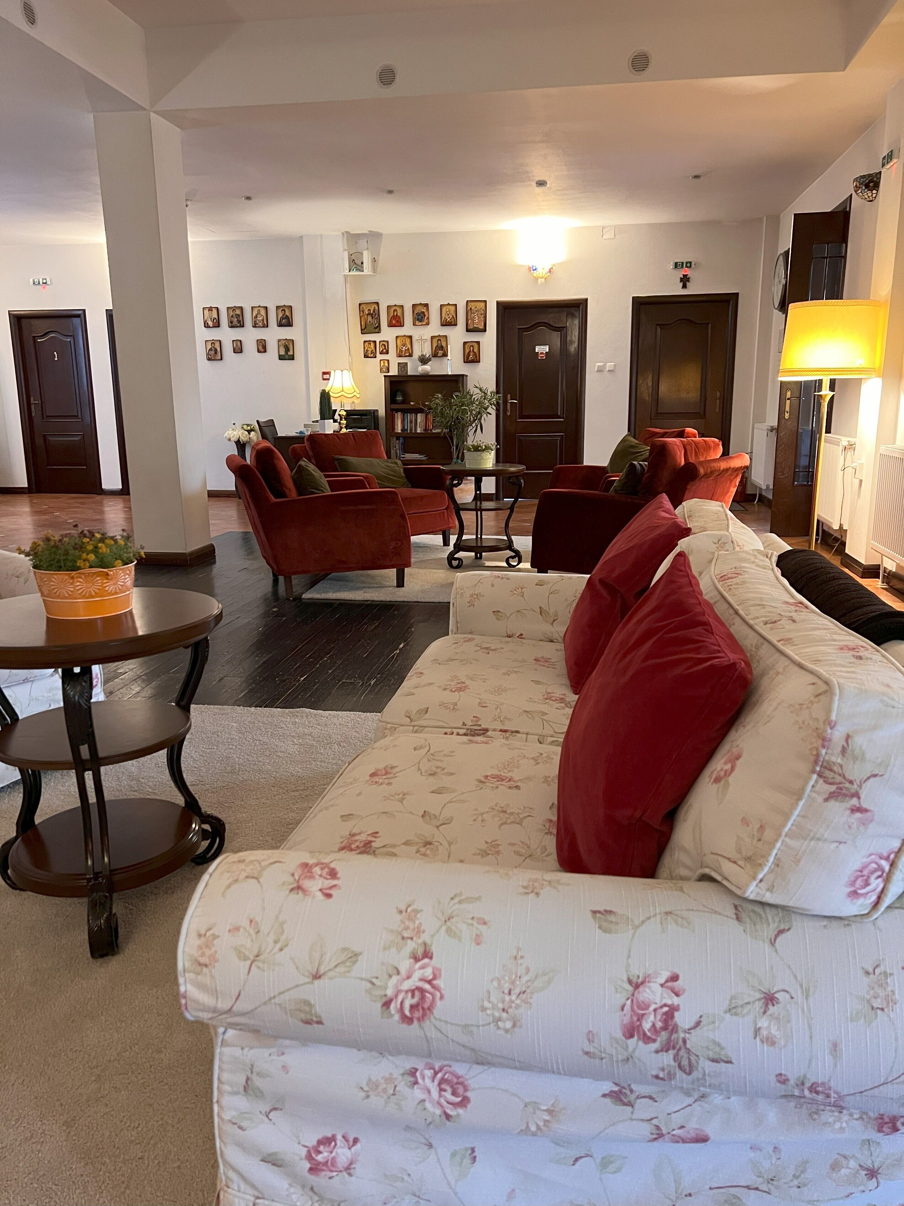 Sala de estar com sofá floral, poltronas vermelhas e uma pequena mesa com uma planta. Quadros emoldurados adornam a parede e uma porta aparece ao fundo.