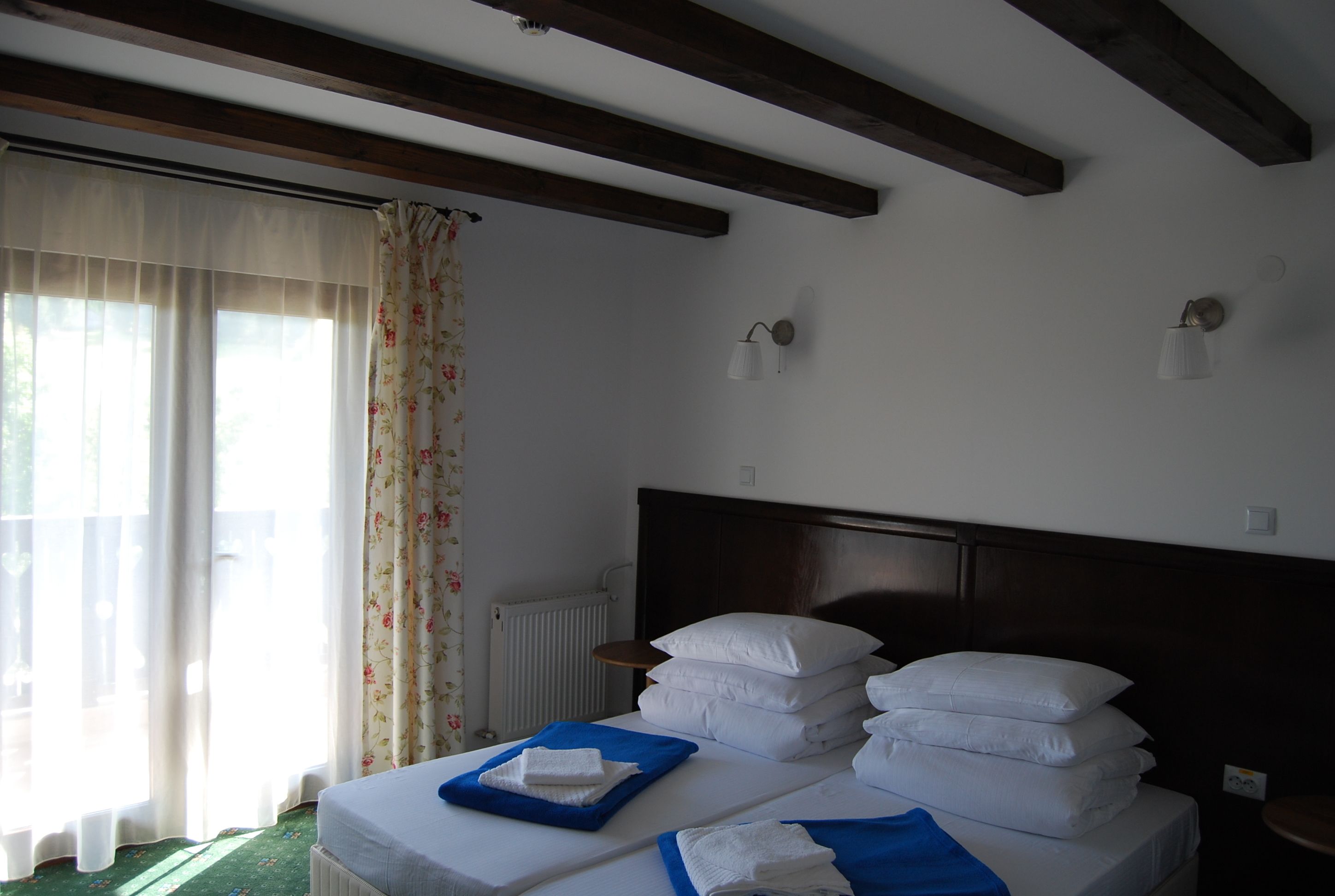 Habitación de hotel con dos camas, cada una con toallas blancas impecables y cubrecamas azules. Hay dos lámparas de pared y una ventana con cortinas estampadas con motivos florales.