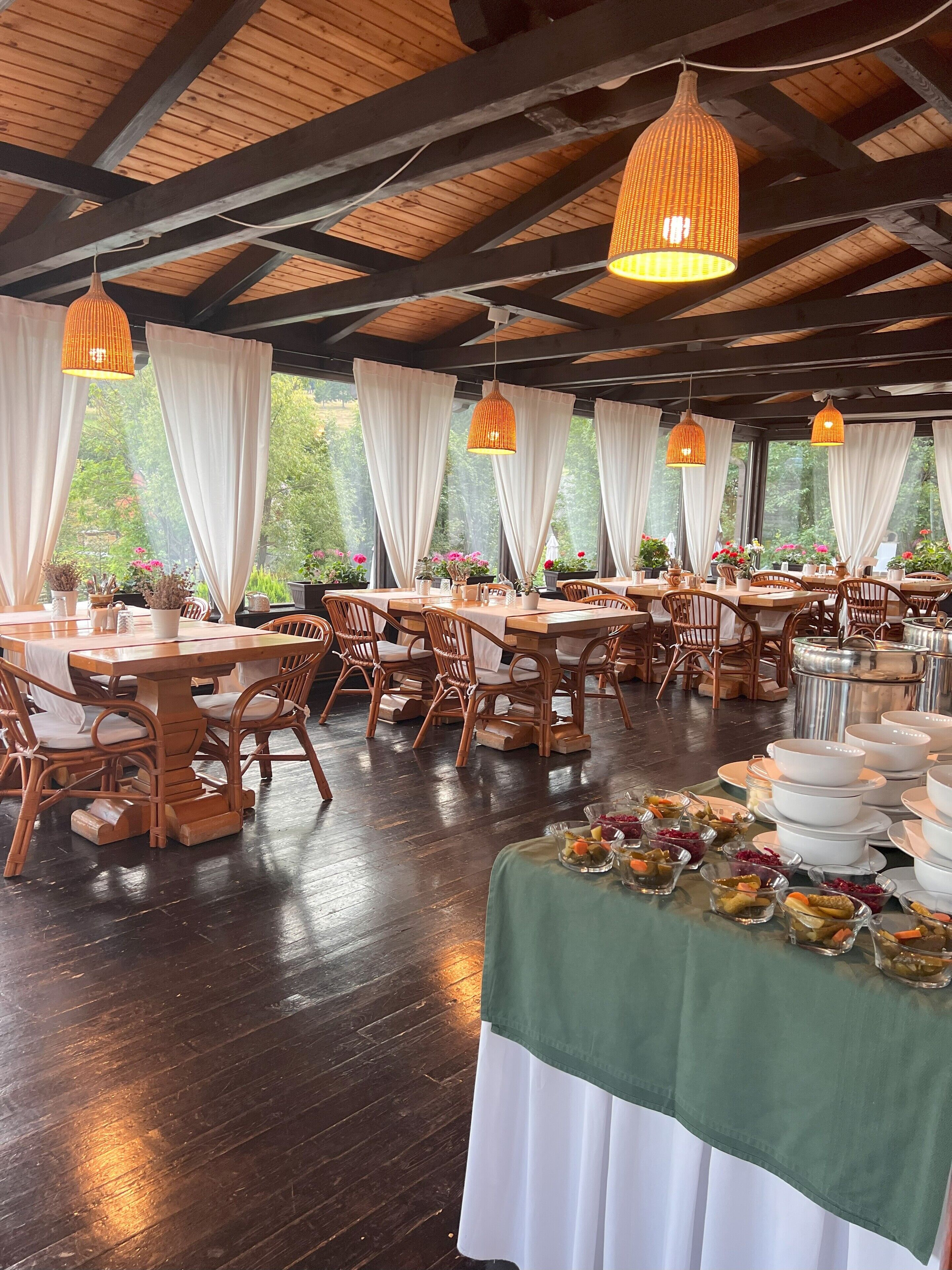 A dining area with wooden tables and chairs, large windows with white curtains, and hanging wicker lamps.