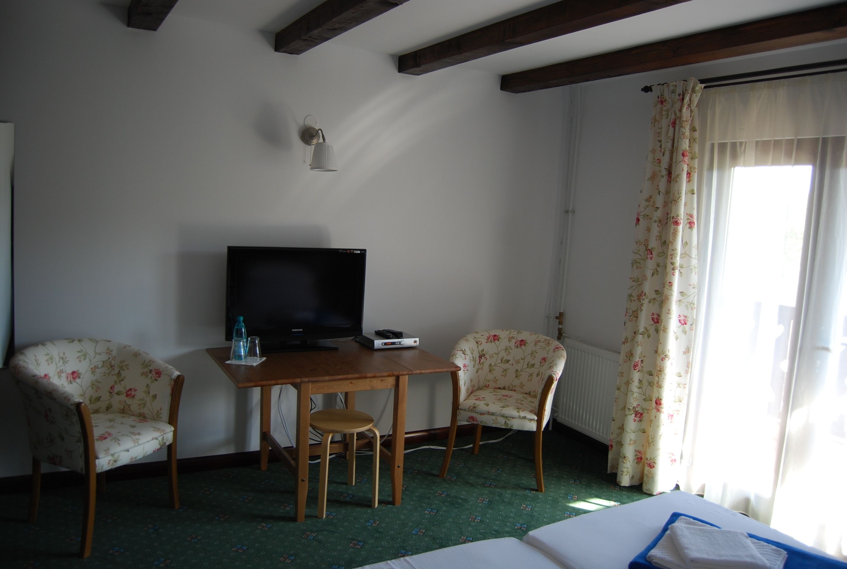 Une chambre d’hôtel avec un lit, un bureau avec une télévision, deux fauteuils à motifs floraux et une fenêtre avec des rideaux.