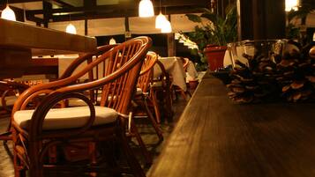A restaurant interior with wooden chairs, a long table, hanging pendant lights, and potted plants.