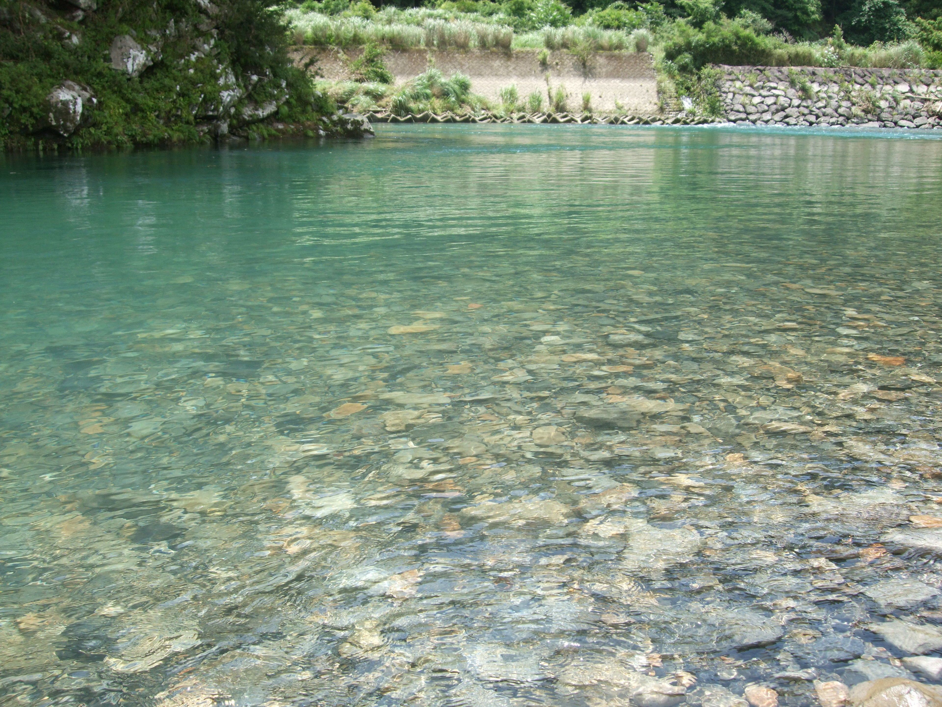 A clear river with visible rocks and a stone wall with vegetation.