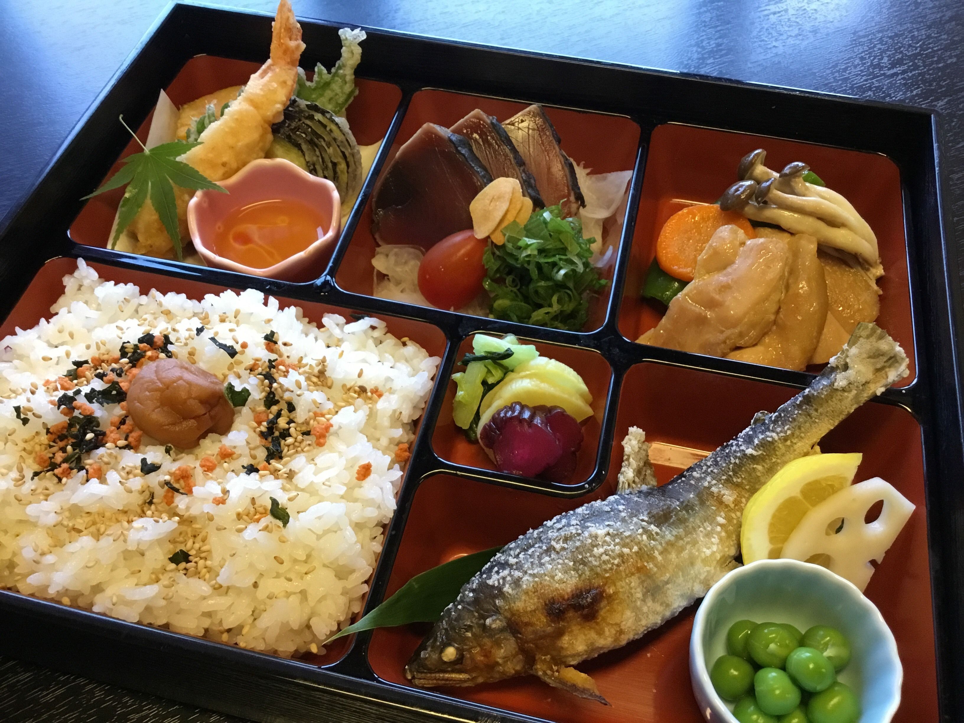 A bento box with rice, fish, vegetables, and a small bowl of green grapes.