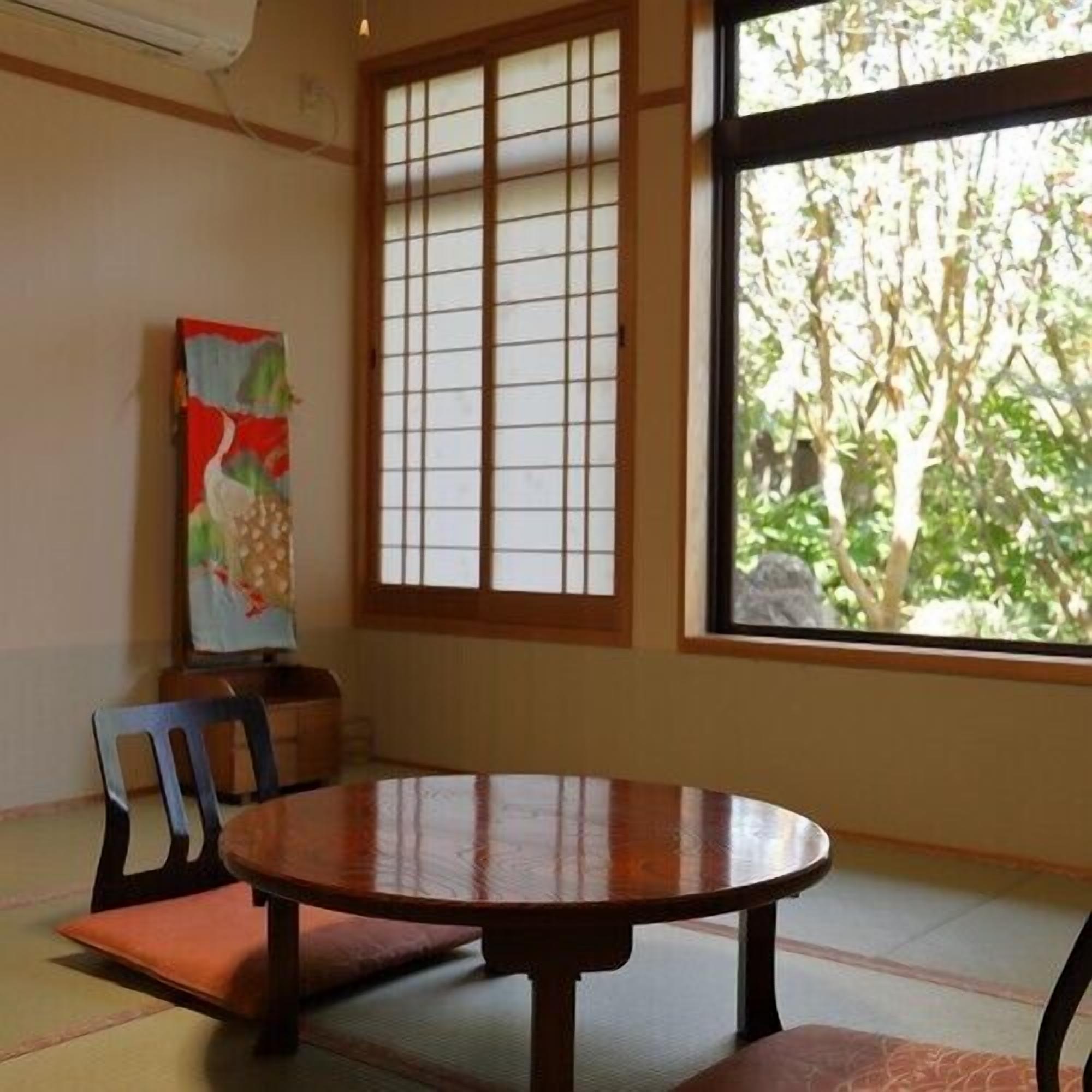 Une pièce avec un sol recouvert de tatamis, une table en bois, une chaise, une fenêtre donnant sur des arbres et un tableau accroché au mur.