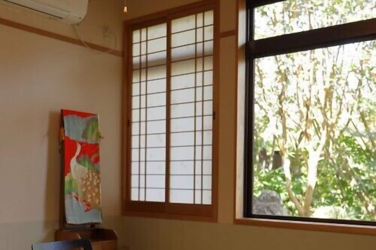 A room with a tatami mat floor, a wooden table, a chair, a window with a view of trees, and a painting on the wall.