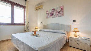 A bedroom with a bed, two bedside tables, a nightstand, a window with curtains, an air conditioning unit, and a floral painting on the wall.