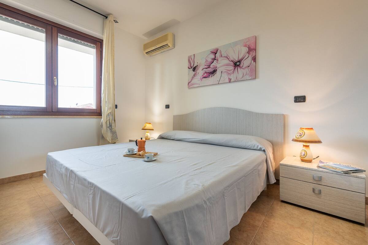 Une chambre à coucher comprenant un lit, deux tables de chevet, une commode, une fenêtre avec des rideaux, un appareil de climatisation et un tableau représentant des motifs floraux accroché au mur.