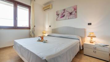 Um quarto com cama, duas mesinhas de cabeceira, um criado-mudo, janela com cortinas, ar condicionado e um quadro com flores na parede.