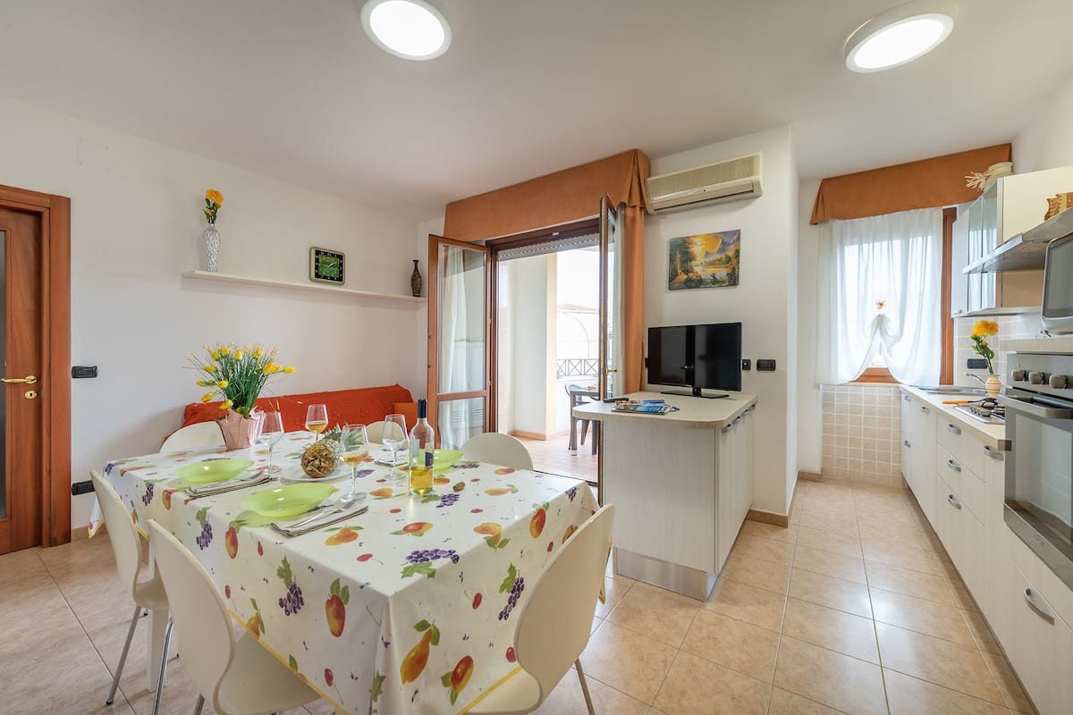 Un coin repas avec une table dressée pour quatre personnes, un téléviseur et un espace cuisine équipé d’une cuisinière et d’un four.