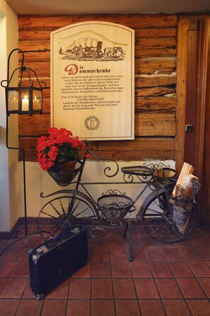 Paredes de madeira rústica com uma placa histórica emoldurada, uma bicicleta antiga com cestas e uma mala preta.