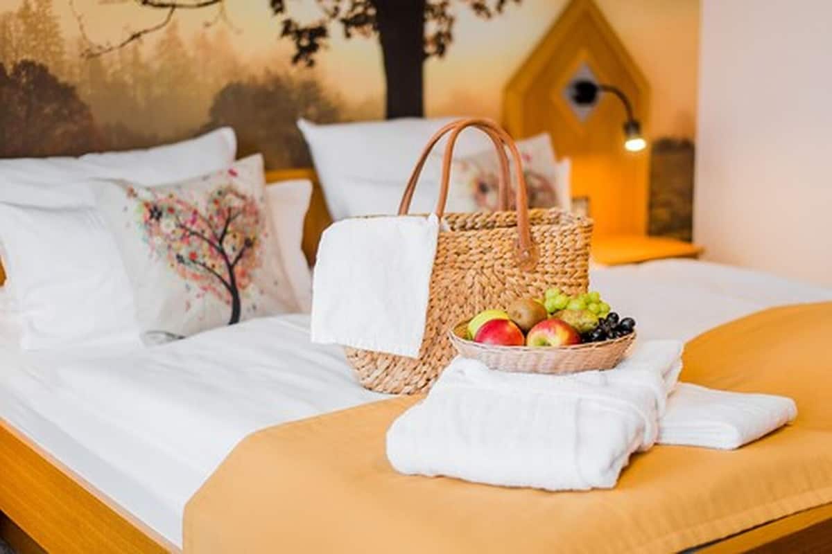 A bed with white linens, a woven basket with fruit, and a white towel on the bed.