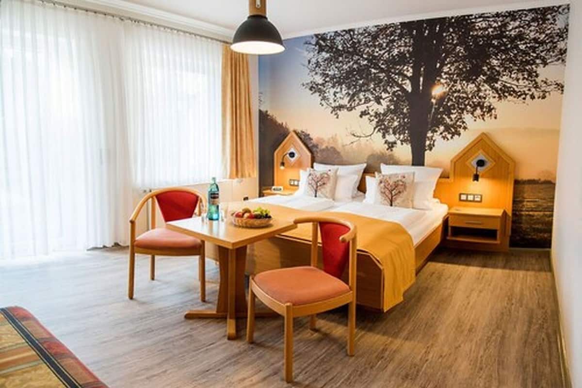 A cozy room with a bed, two chairs, a table with a fruit bowl, a bottle, and a wall mural of trees.