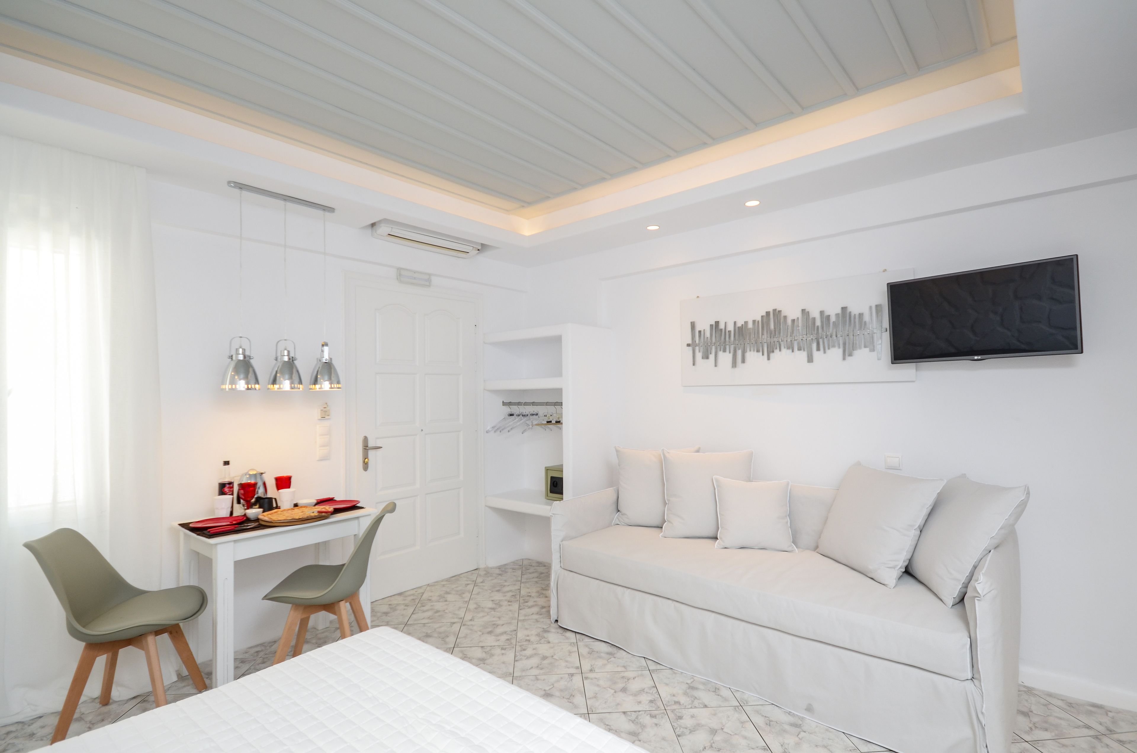 A modern, well-lit room with a white sofa, two chairs, a dining table, and a flat-screen TV mounted on the wall.