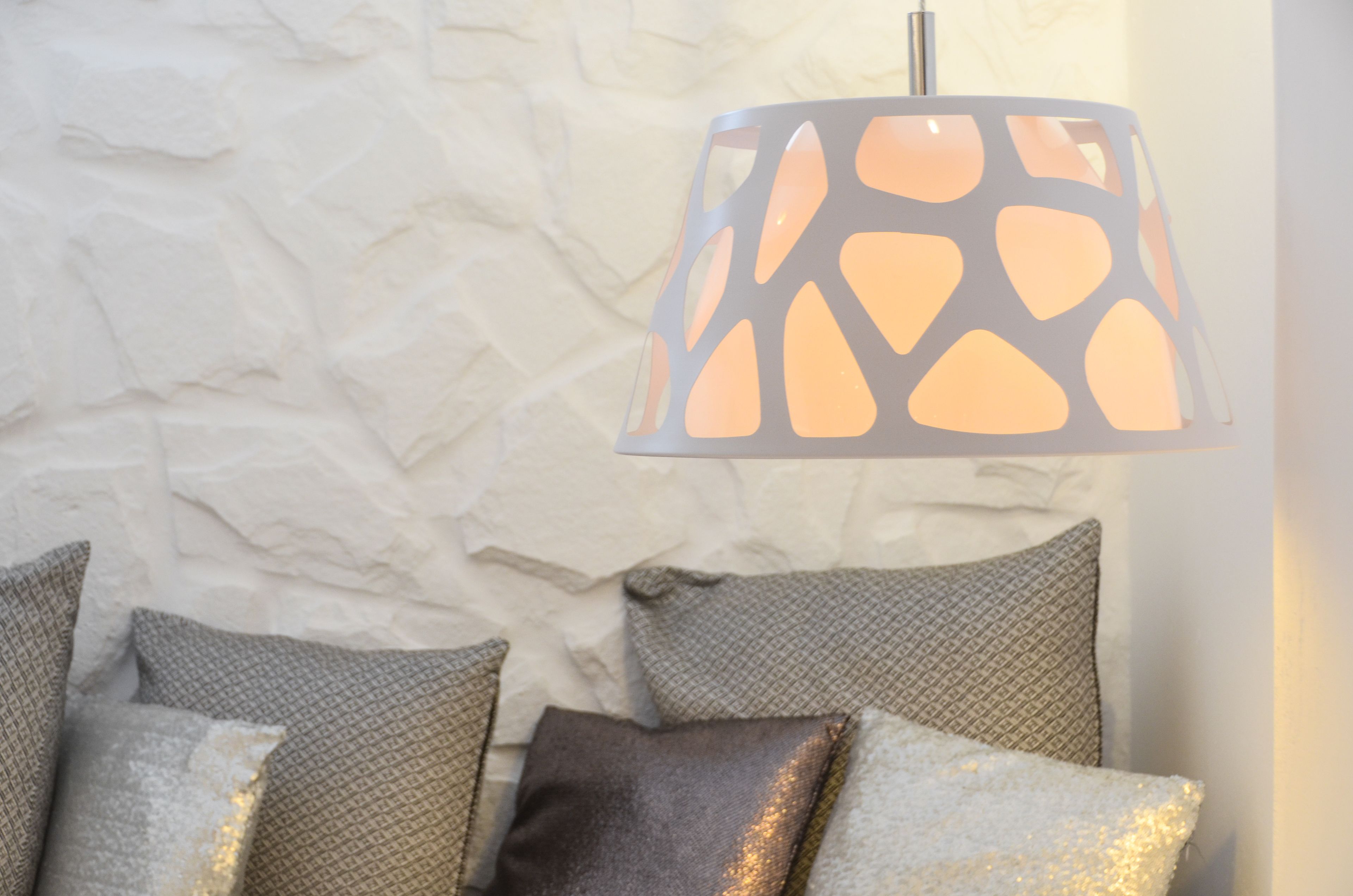 Eine Lampe mit geometrischem Muster, eine strukturierte Wand und ein Bett mit gemusterten Kissen.