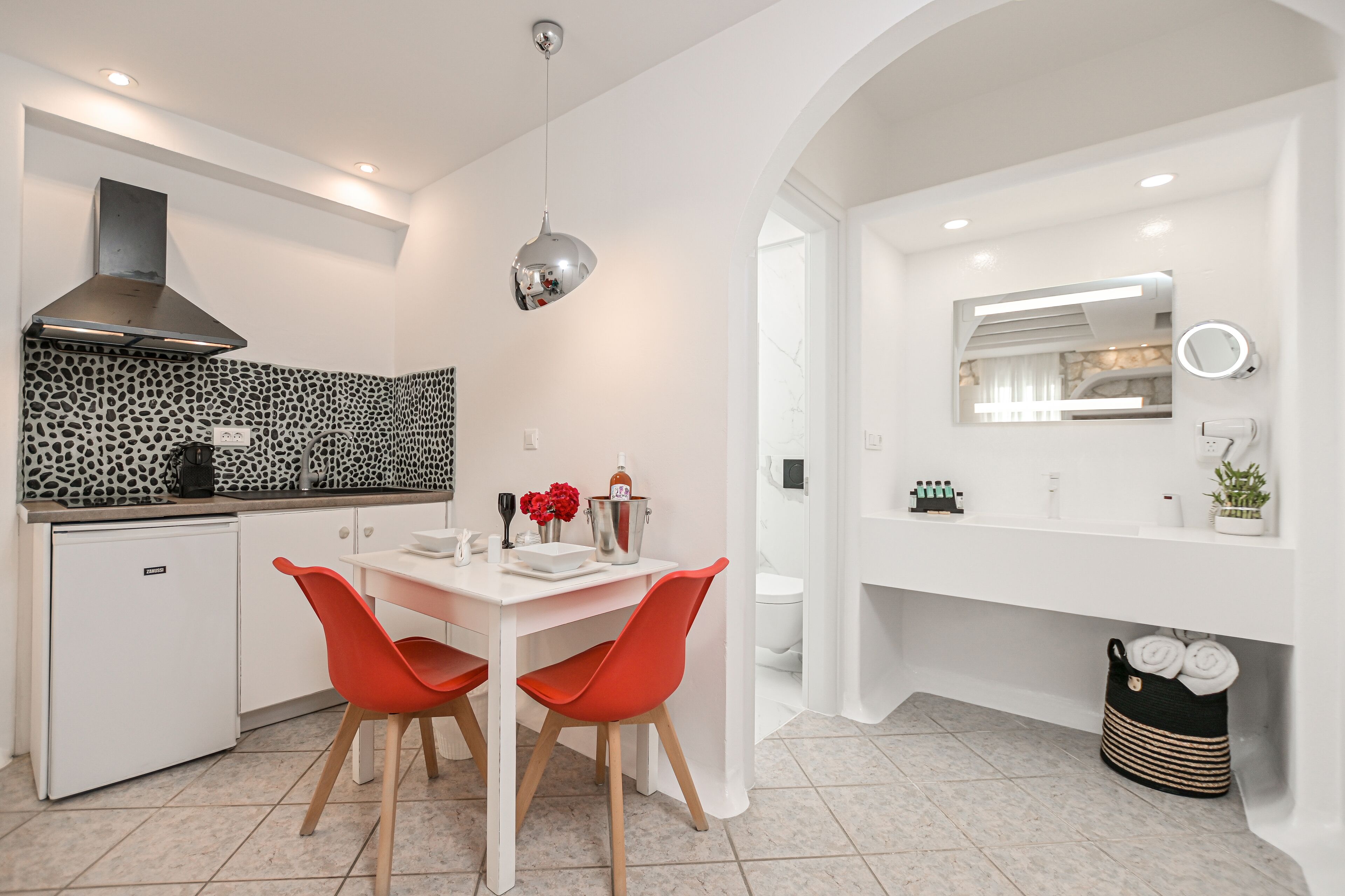 Una cocina moderna con encimera blanca, horno integrado y un salpicadero con diseño. Se ve una mesa de comedor con dos sillas rojas y una lámpara colgante encima.