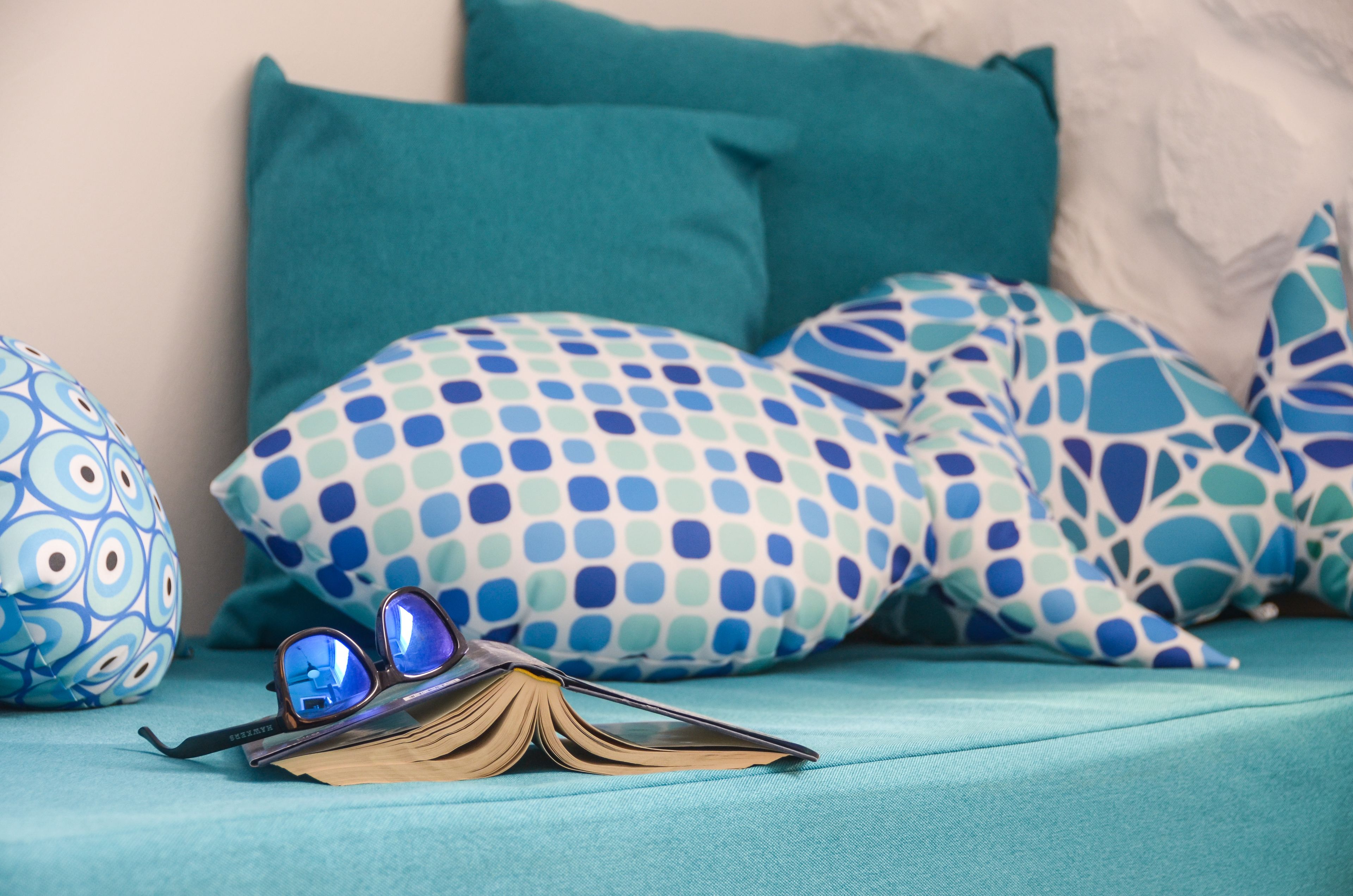 A bed with teal bedding and patterned pillows, an open book, and a pair of glasses on the bed.