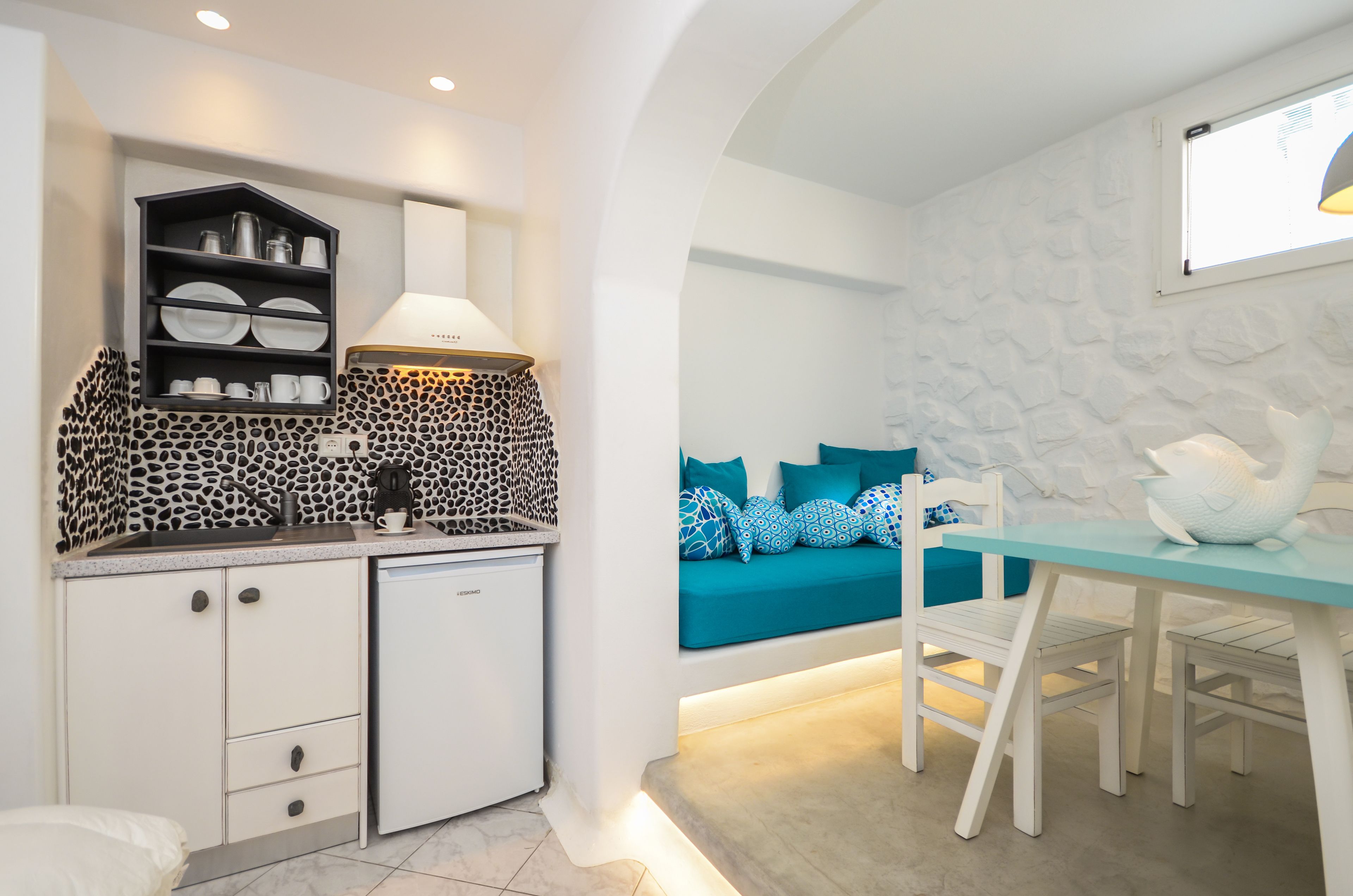 A compact kitchen with a built-in stove, a coffee machine, and a dishwasher. There is a seating area with a teal sofa and patterned cushions.