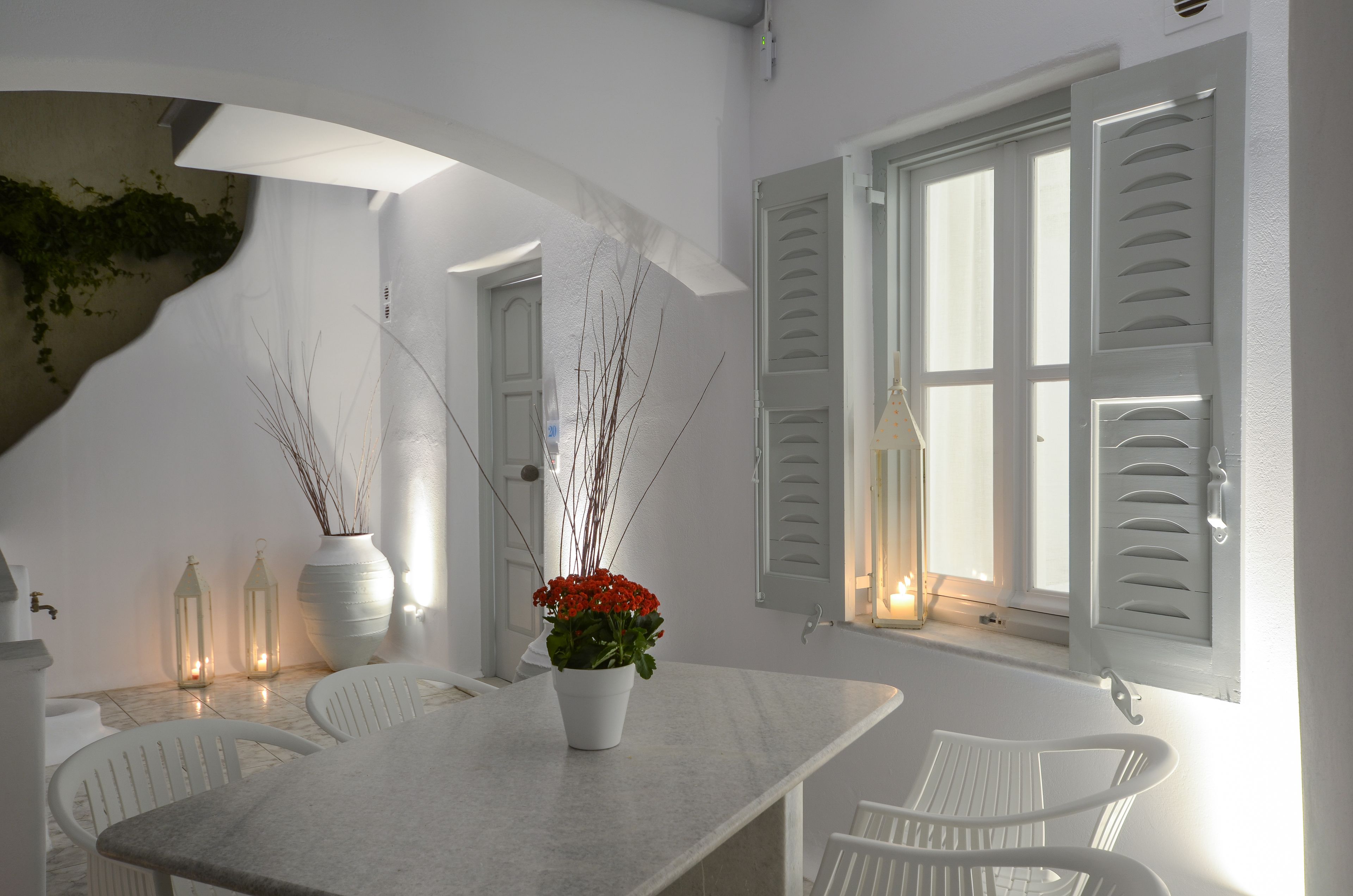 A dining area with a white table, chairs, and a vase with red flowers. There are two lanterns with candles on the table.