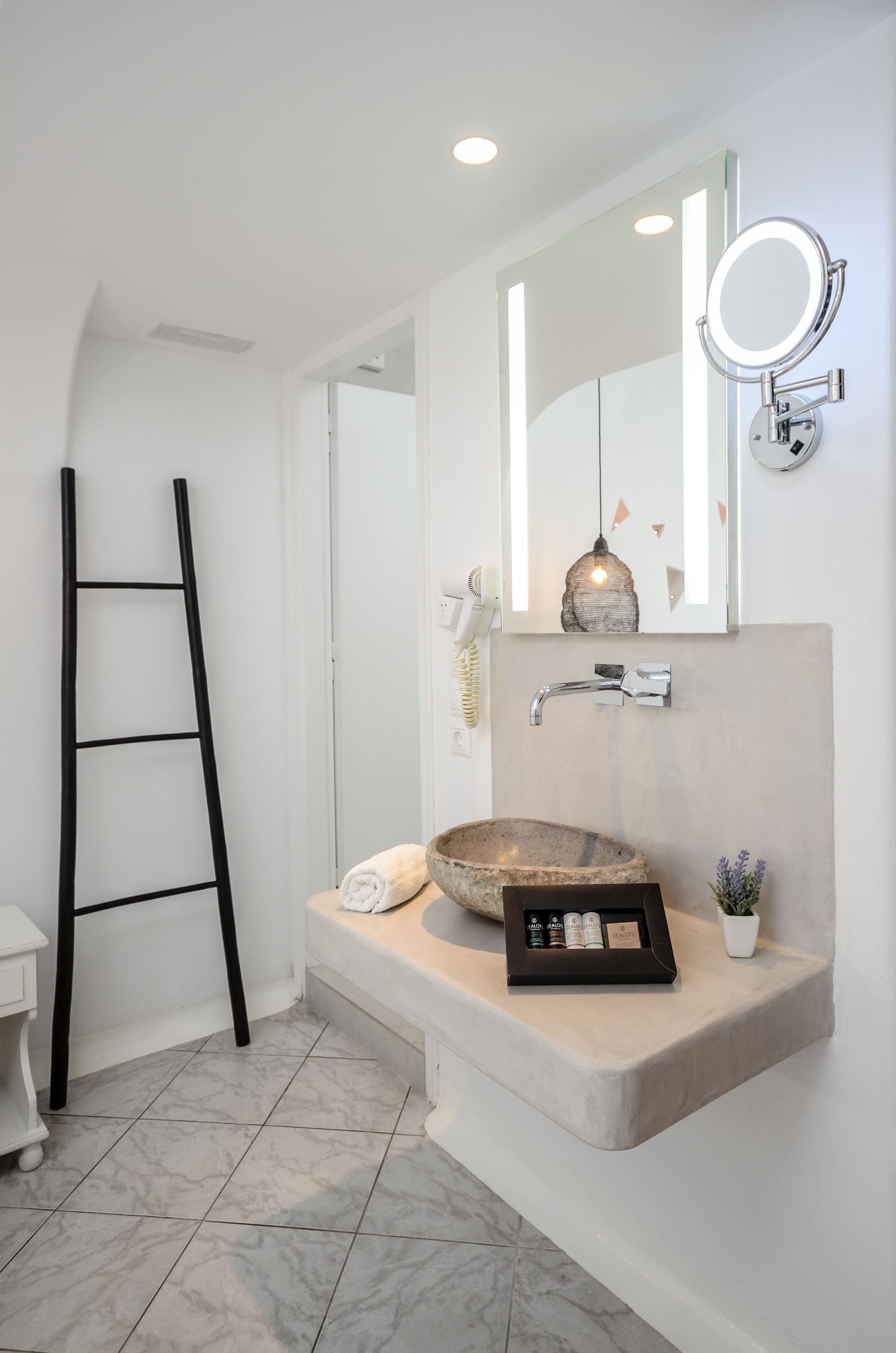 A bathroom with a marble floor, a beige sink with a large mirror above it, a black ladder, and a small potted plant.