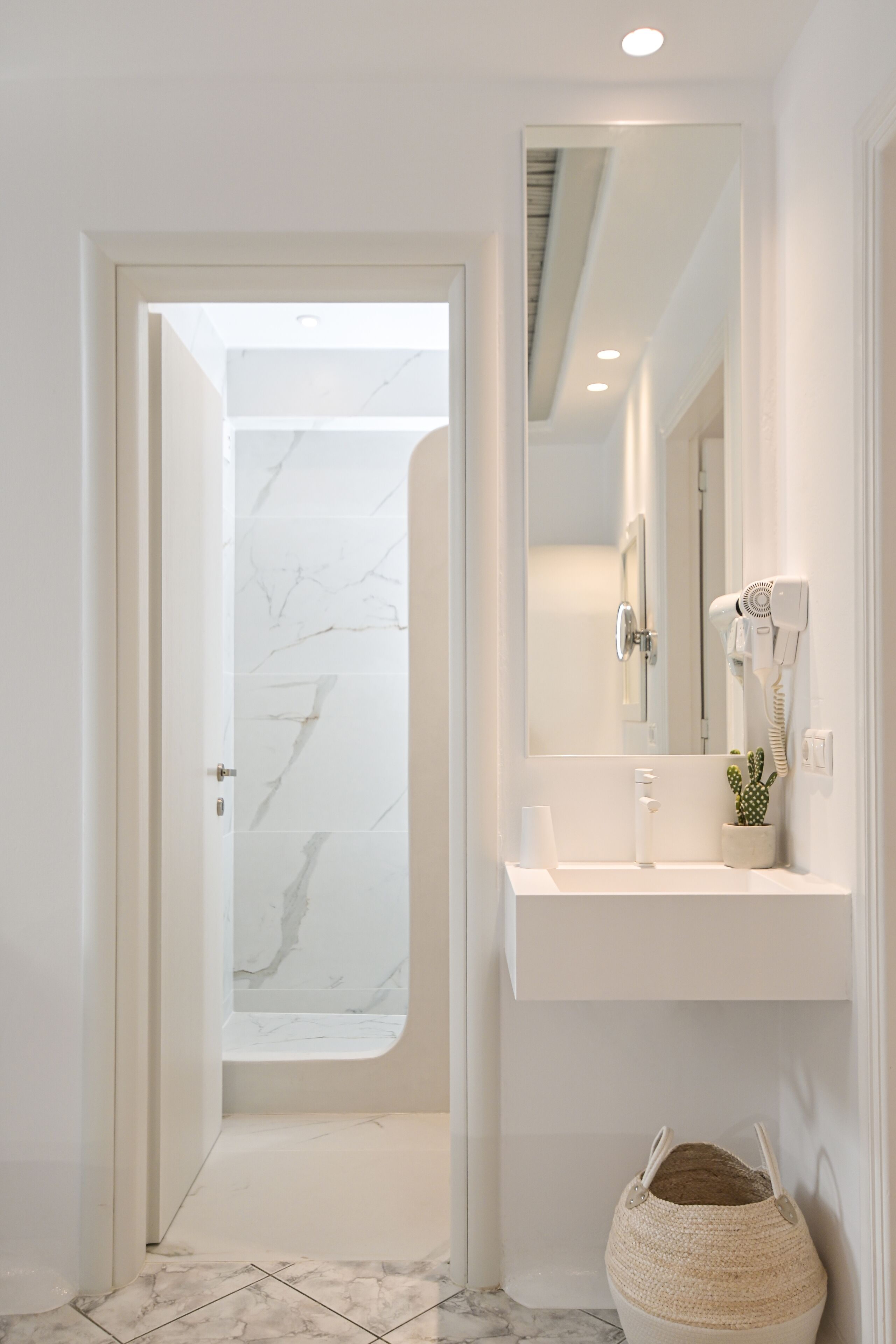 A modern bathroom with a white marble shower, a white sink with a hairdryer, and a small potted cactus.