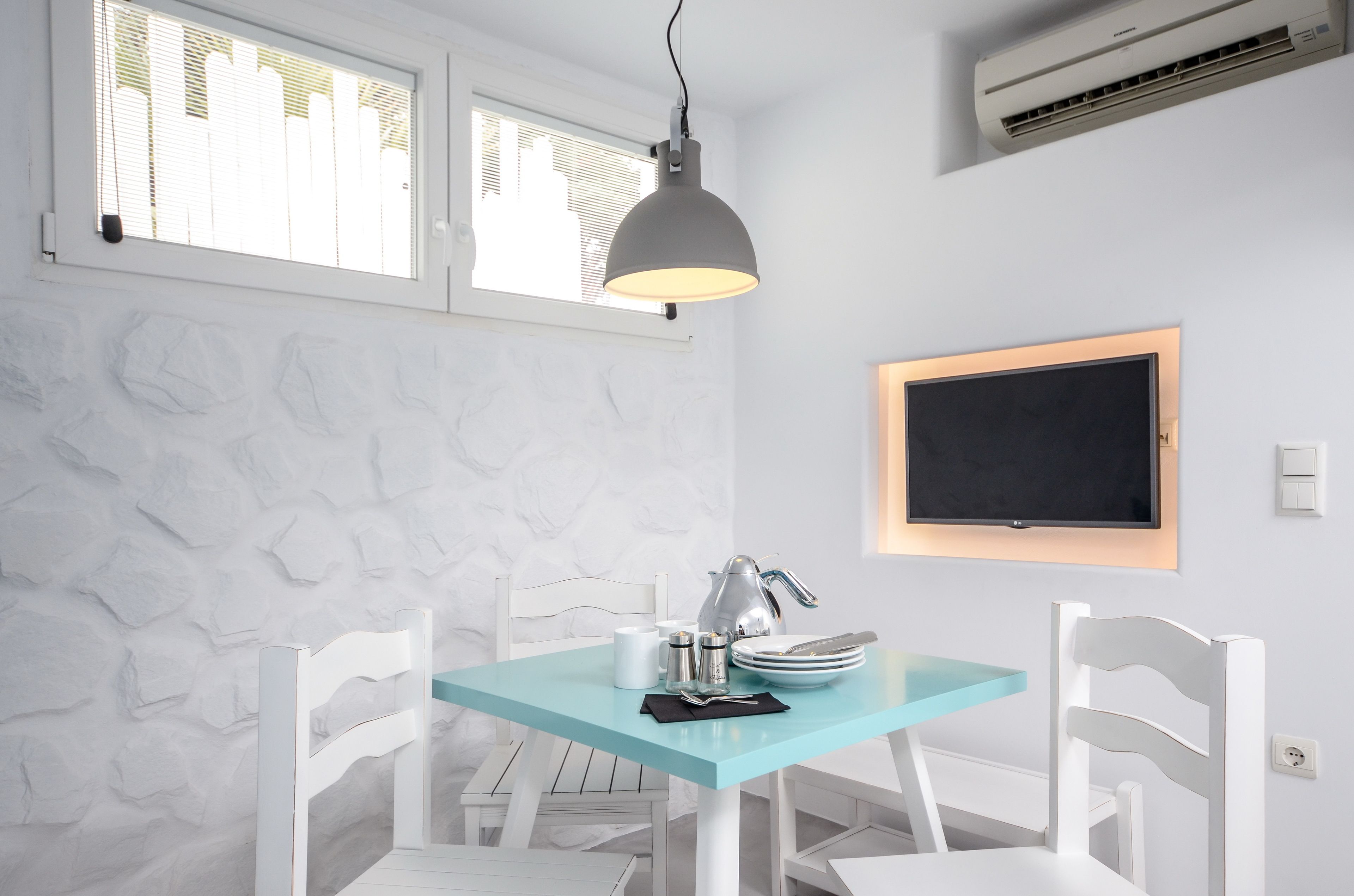 A dining area with a light blue table, white chairs, a hanging pendant light, a wall-mounted TV, and a stone-textured wall.