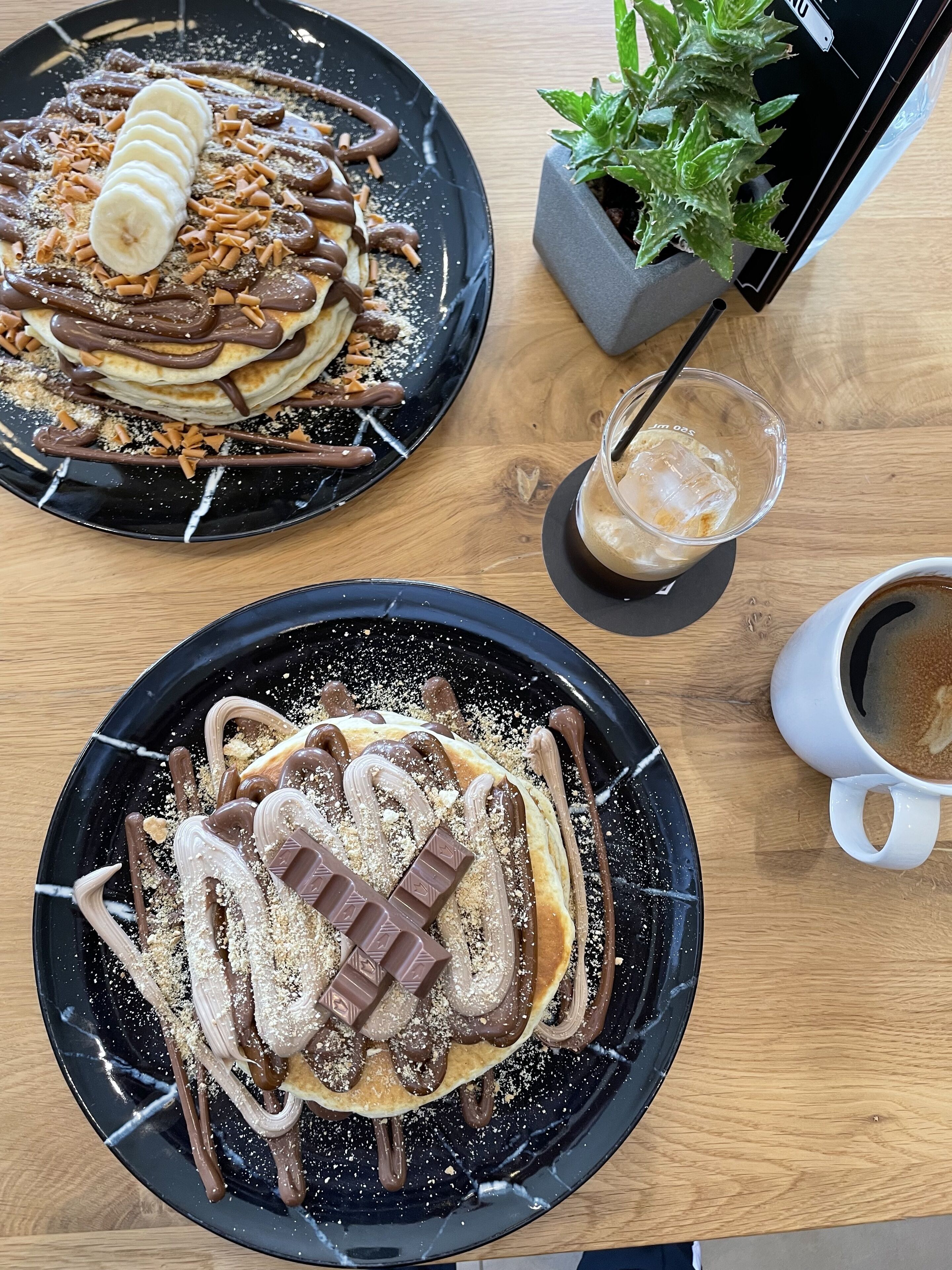 Zwei Dessertteller mit Schokoladen- und Bananenbelegen, ein Glas Eiskaffee und eine Tasse Kaffee auf einem Holztisch.