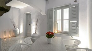 Comedor blanco con una mesa, sillas y un jarrón de flores rojas. Hay dos plantas en macetas y una ventana con contraventanas.