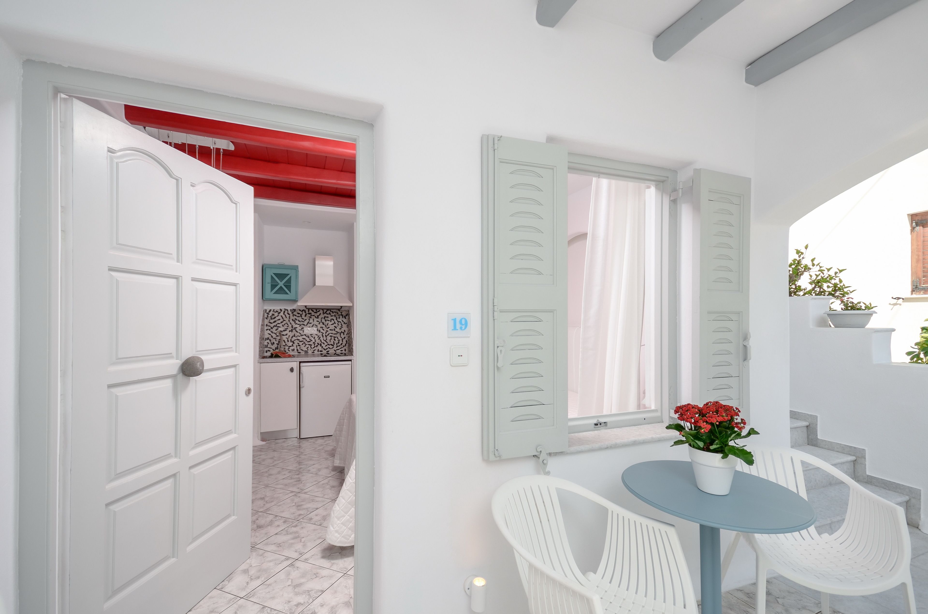 A room with a white door, a window with shutters, a small table with a potted plant, and white chairs.
