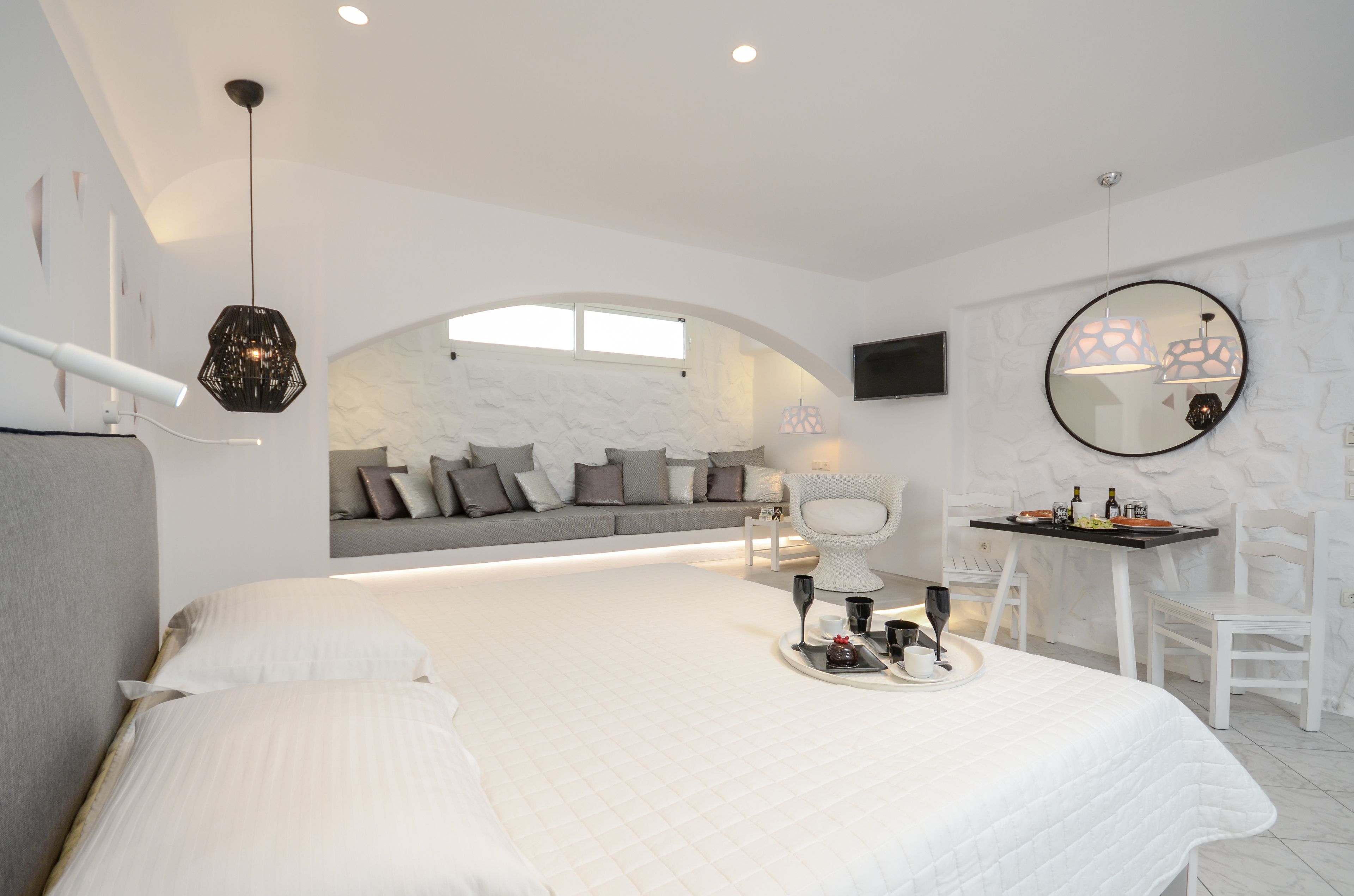 A modern bedroom with a large bed, a seating area with a sofa and chairs, a dining table with a tray of items, and a round mirror on the wall.