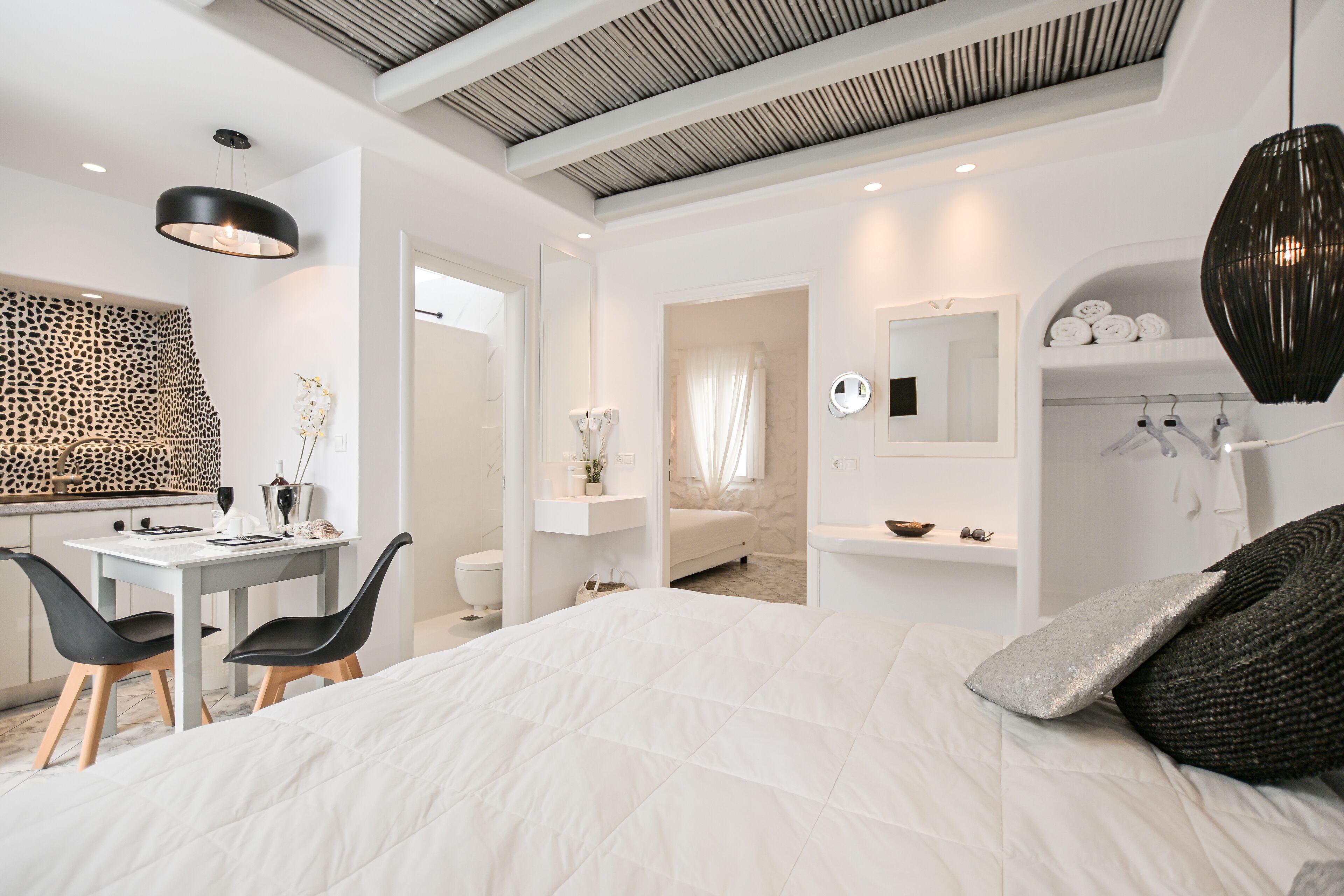 A modern bedroom with a large bed, a desk with chairs, a bathroom, and a kitchenette.