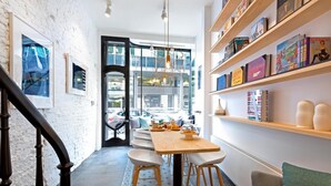 A modern kitchen with a wooden table and bar stools, a staircase with dark wooden balusters, and a view of the street through large windows. - HAPPY Guesthouse (Brussels)