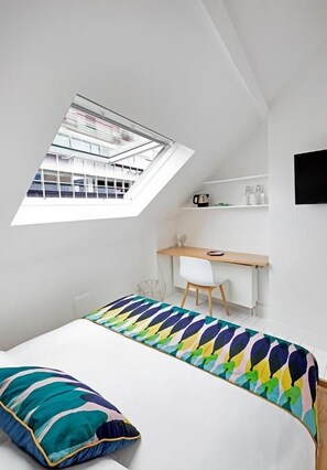 A bedroom with a skylight, a bed with a colorful blanket, a desk with a chair, and a shelf with items. - HAPPY Guesthouse (Brussels)