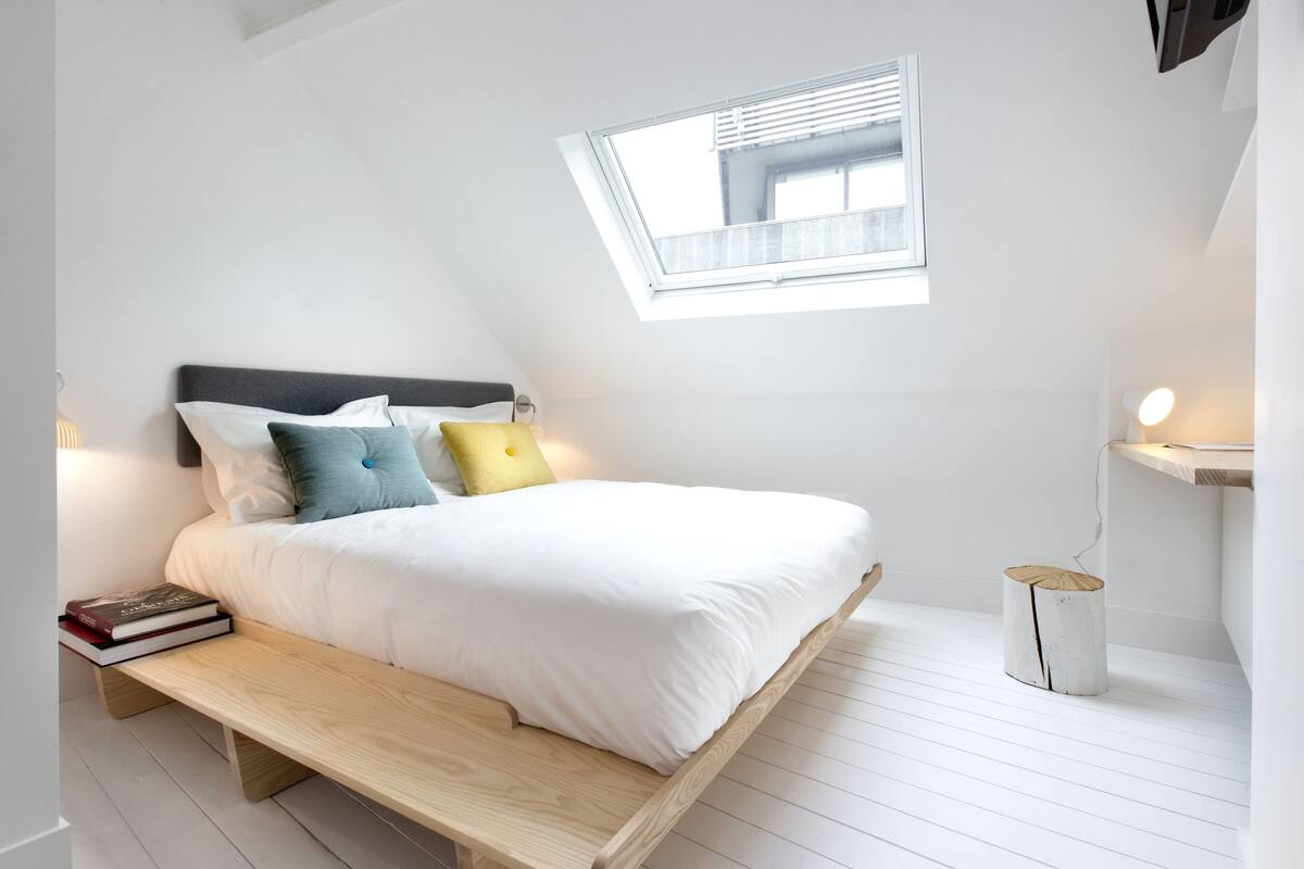 A bedroom with a sloped ceiling, a skylight, a bed with white bedding, a bedside table with books, and a bedside lamp.
