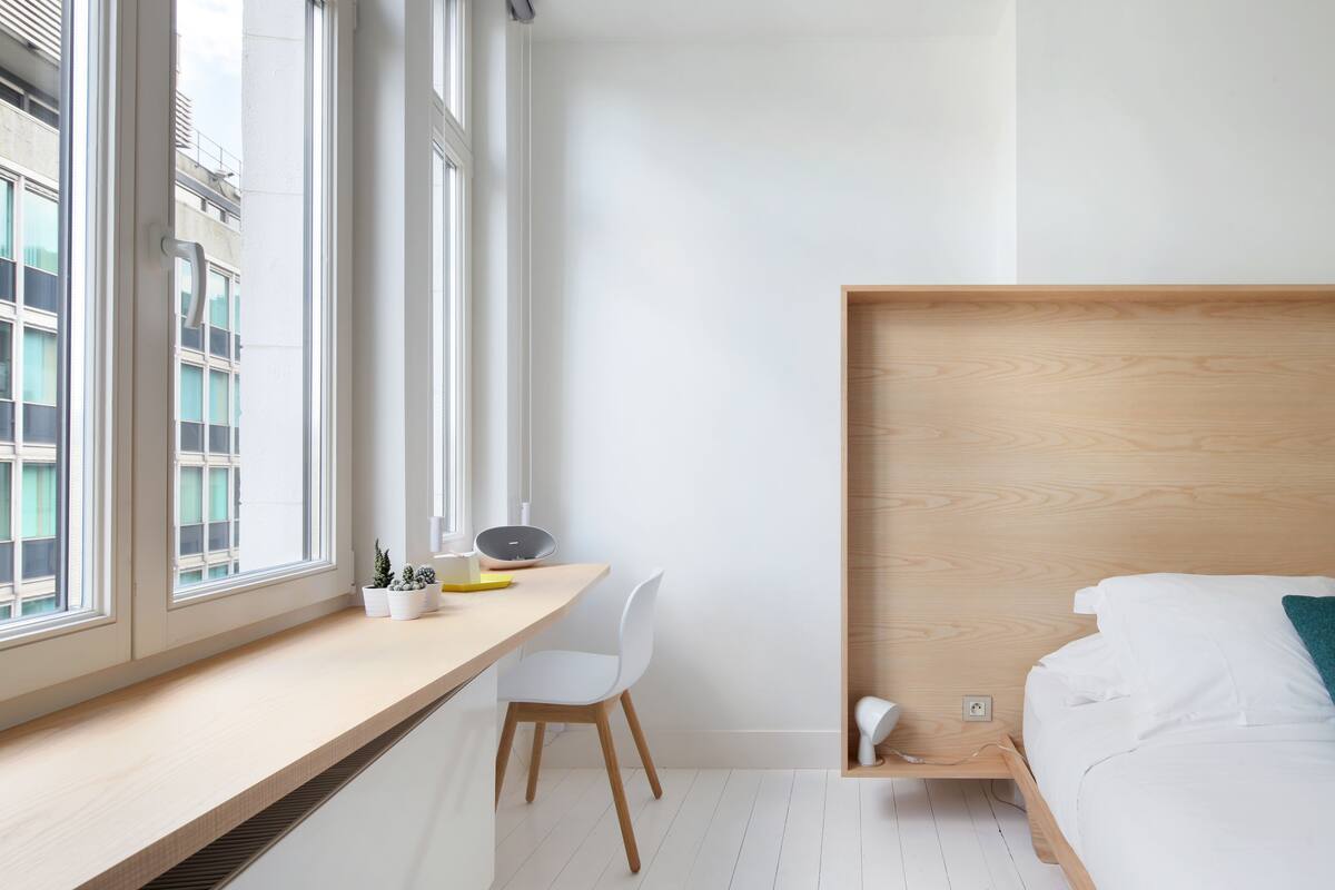 A modern bedroom with a wooden headboard, a bedside table, a chair, and a desk with a laptop and plants.