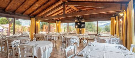 Un restaurante con vigas de madera, mesas redondas, sillas blancas y ventanas amplias con cortinas.