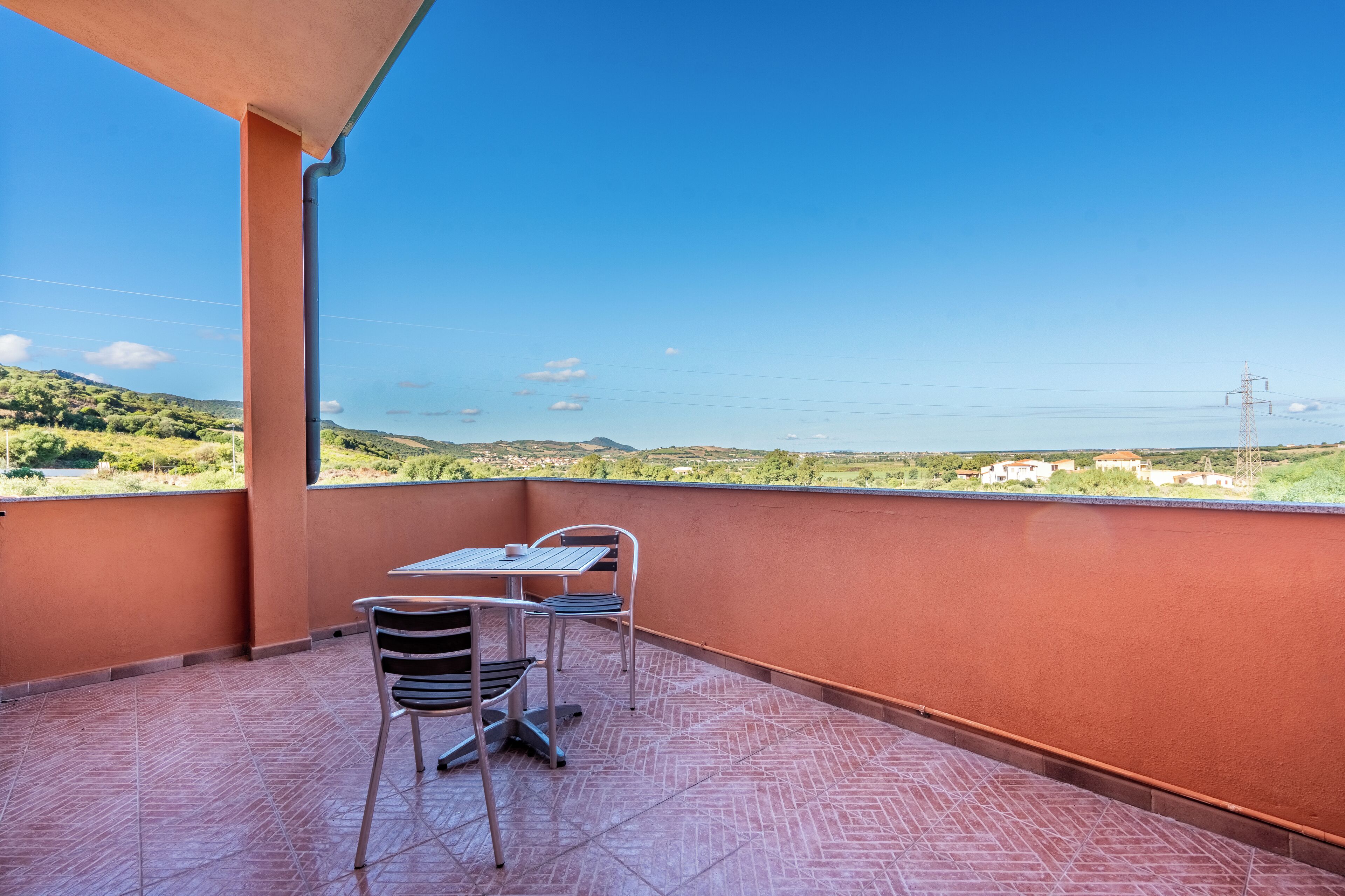 Un balcón con mesa y sillas, con vistas a un paisaje pintoresco con colinas y una lejana vista de agua.