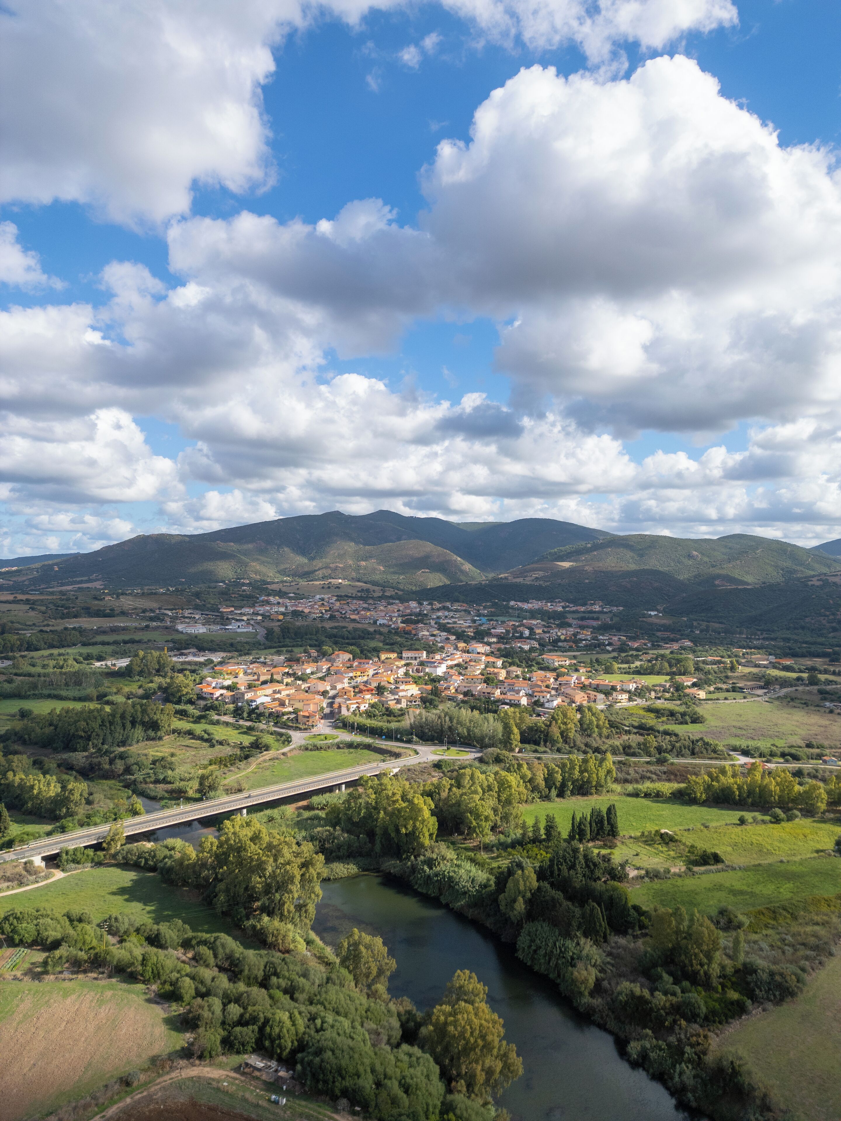 A panoramic view of a town with a river, green fields, and mountains in the background.