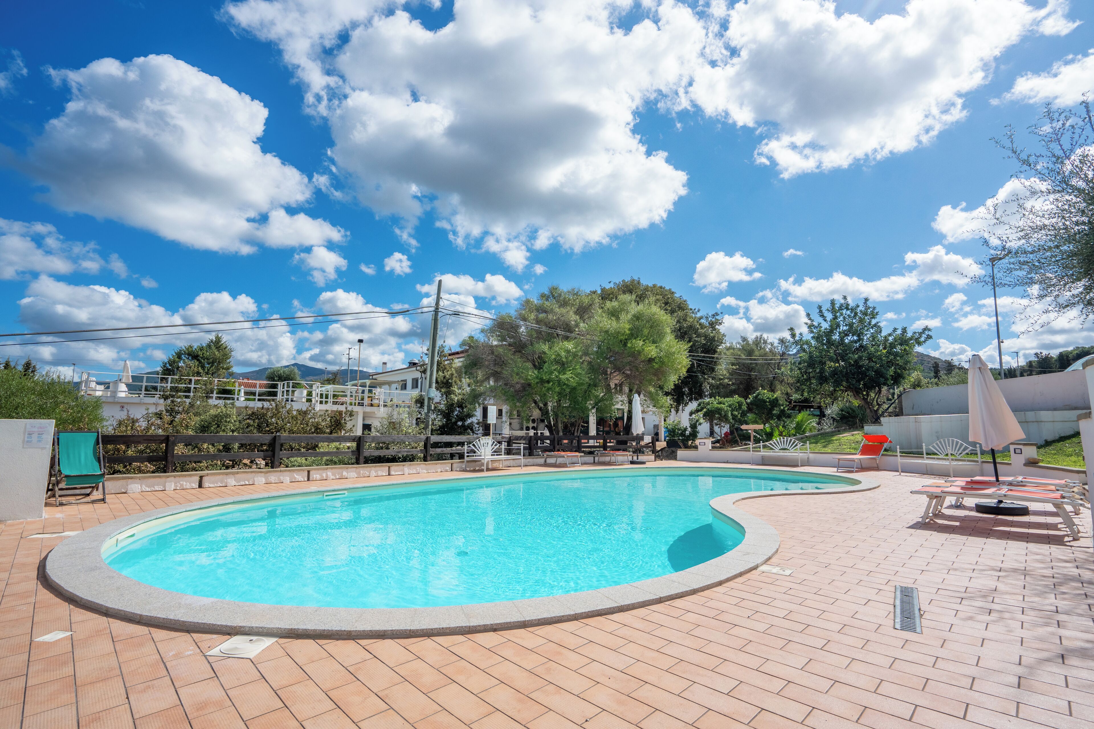 A swimming pool with a tiled deck, surrounded by lounge chairs and umbrellas, under a partly cloudy sky.