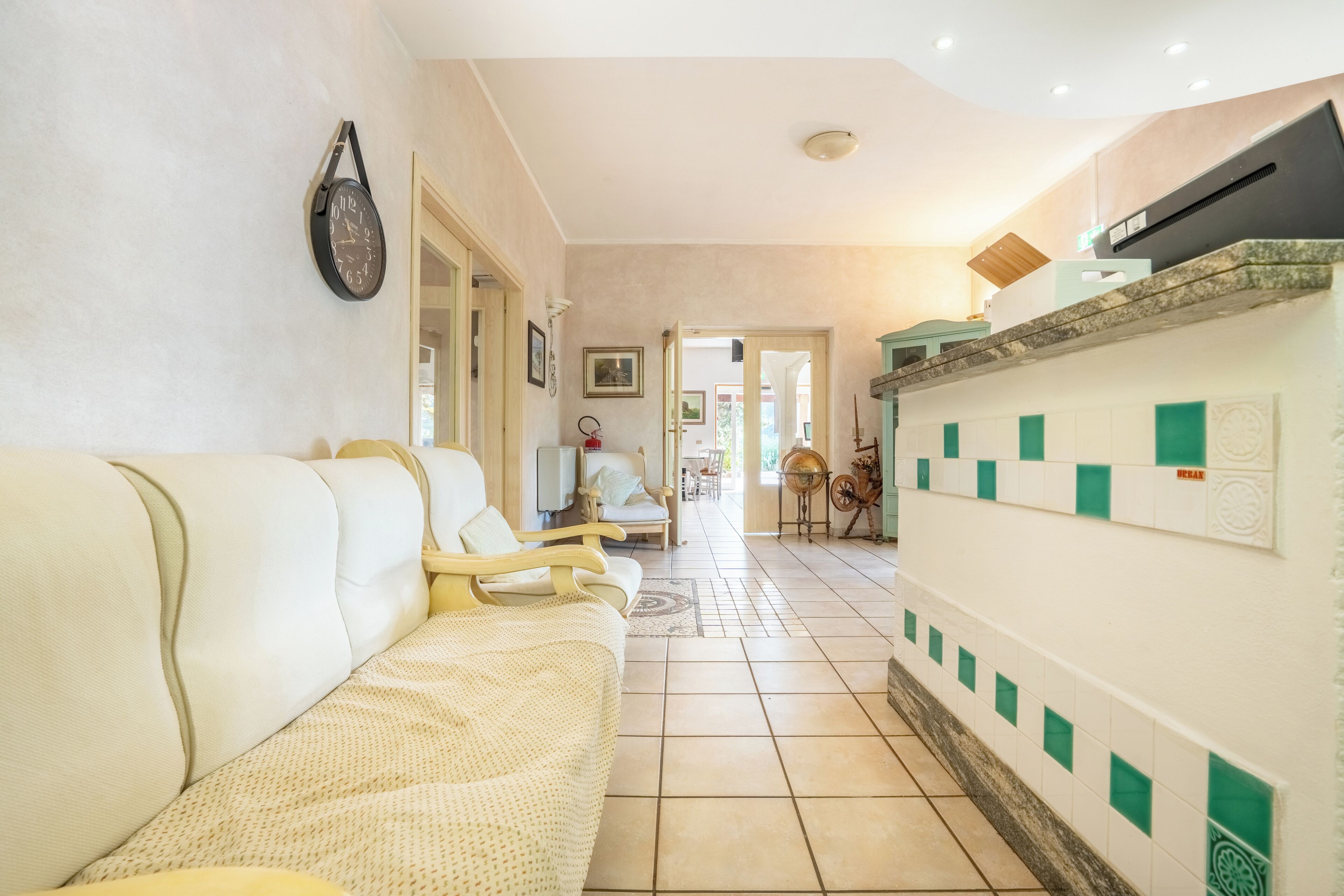 A hallway with a sofa, a wall clock, and a kitchen area with green and white tiles.