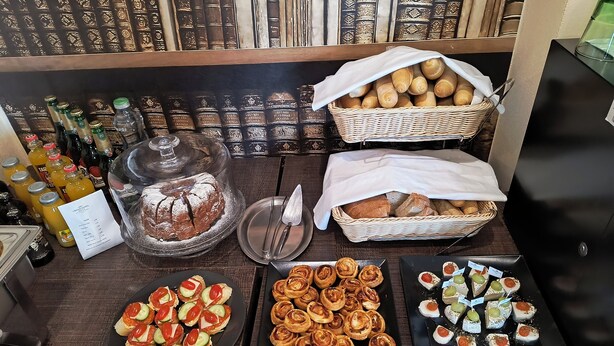A buffet table with various food items, including bread, pastries, and a cake, along with bottles of beverages. - Grand Hotel Senica (Senica)