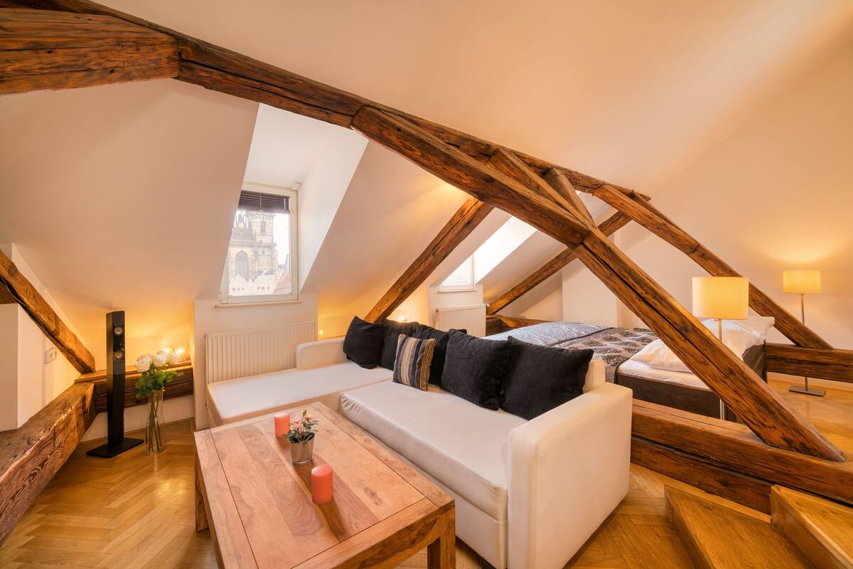 Un dormitorio acogedor en el ático con techo de vigas de madera, un sofá blanco, una mesa de madera y una ventana con vistas a un edificio.