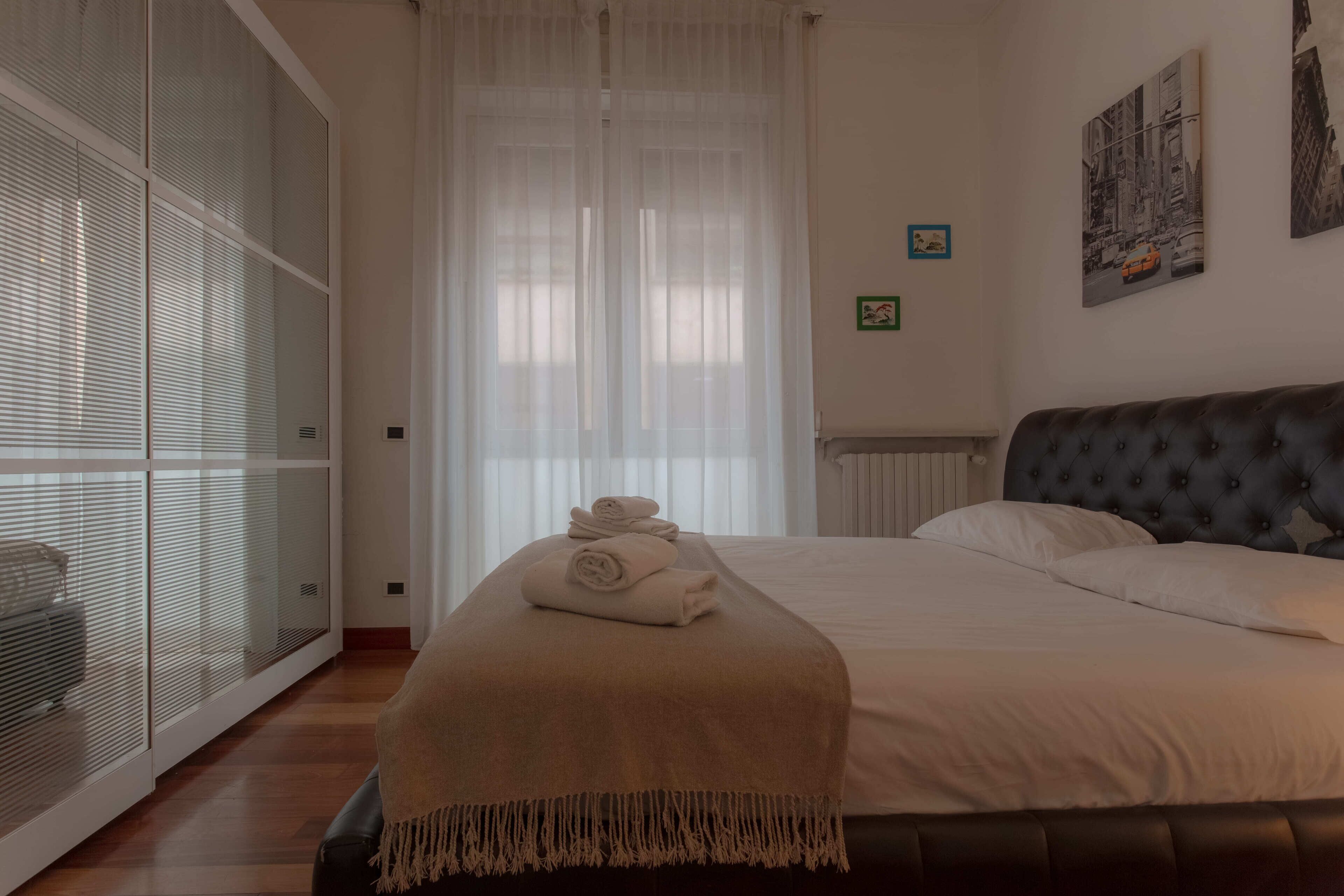 A bedroom with a bed, a large window with blinds, a radiator, and a wall with two framed pictures.