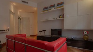 Un salon moderne avec un canapé rouge, un meuble de rangement noir et un téléviseur. On y trouve des meubles blancs et une étagère avec des livres et des objets décoratifs.