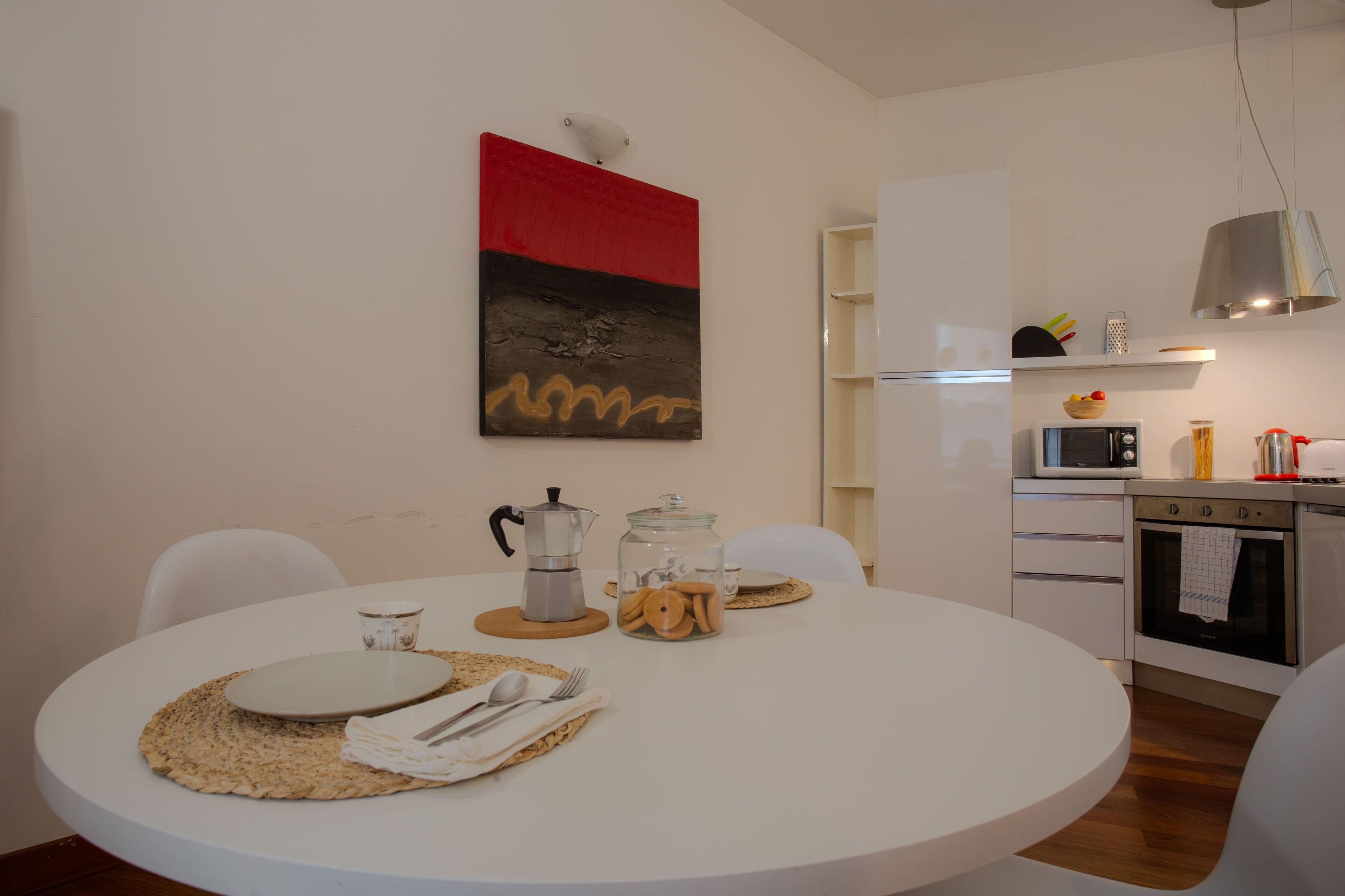 A modern kitchen with white cabinets, a microwave, and a stove. There is a dining table with a placemat, a coffee pot, and a glass jar with cookies.