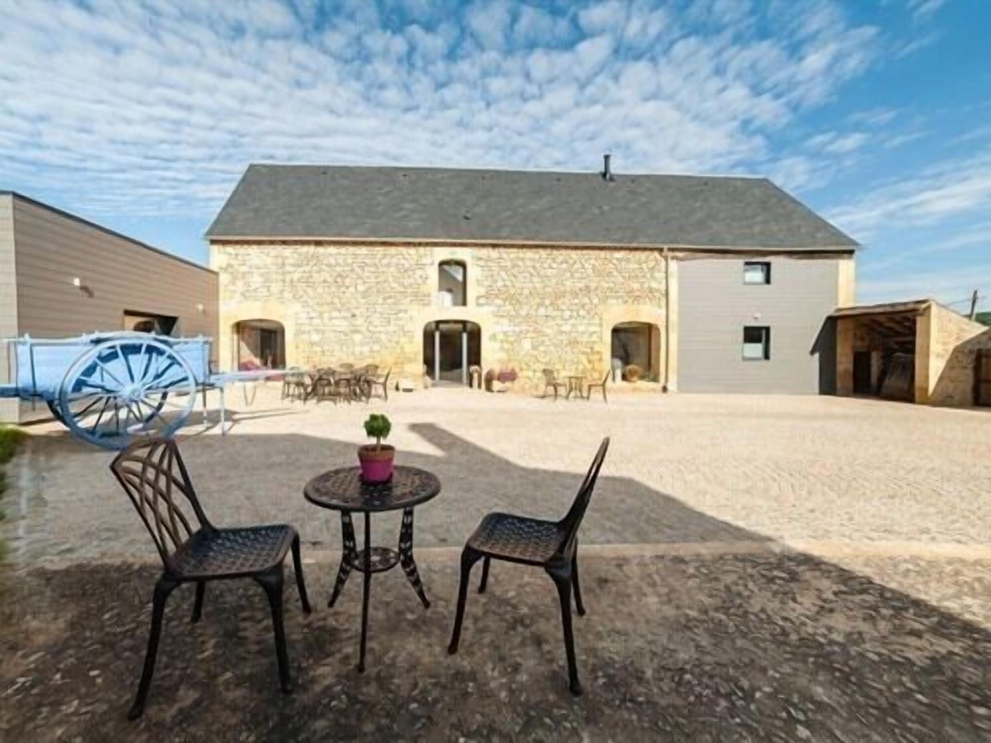 A stone building with a grey roof, a courtyard with a table and chairs, and a blue carriage wheel.
