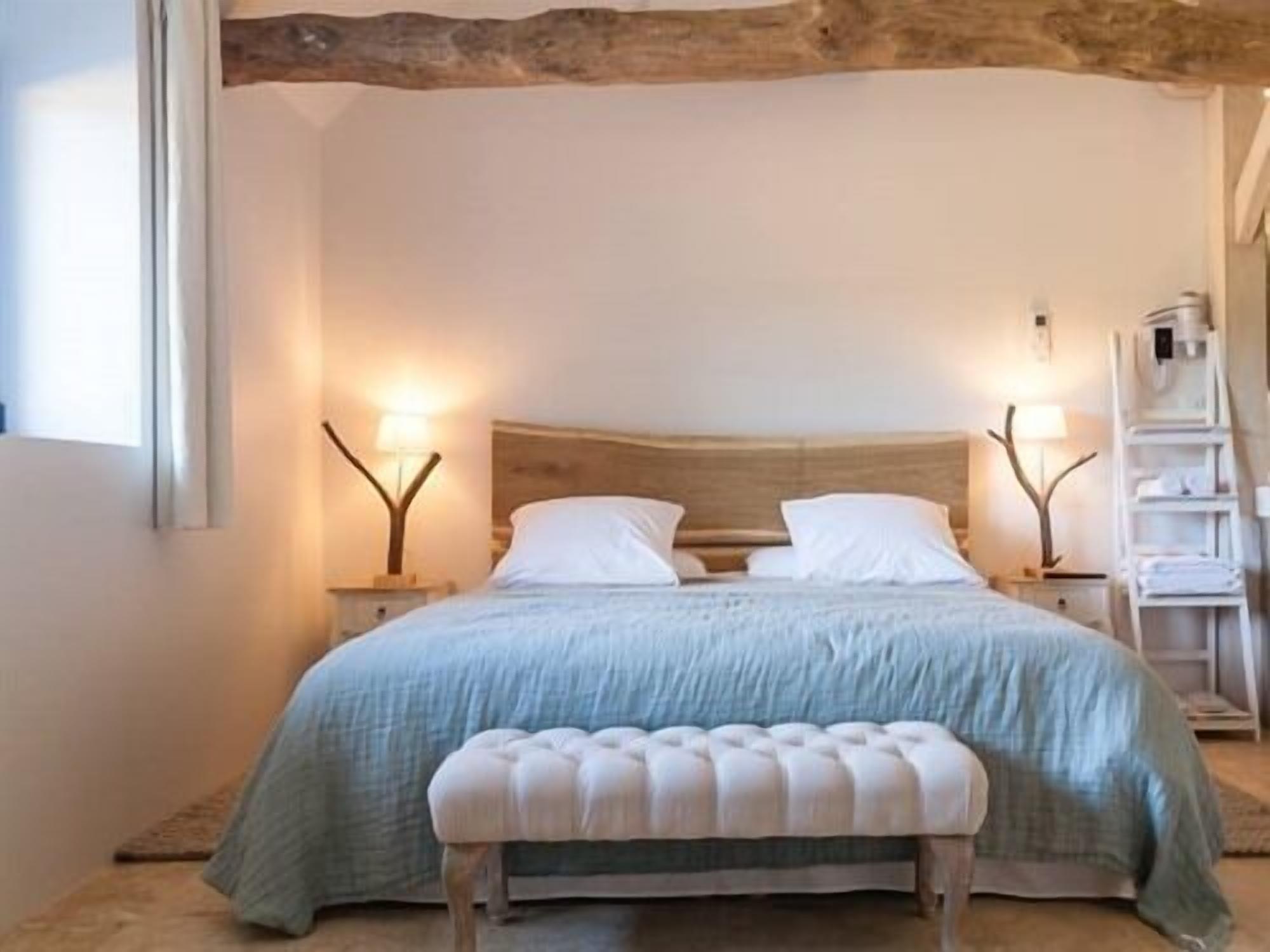 Ein Schlafzimmer mit einem hölzernen Kopfende, einem Bett mit einer hellblauen Bettdecke, zwei Nachttischlampen mit verzweigtem Gestell und einer weißen Bank vor dem Bett.
