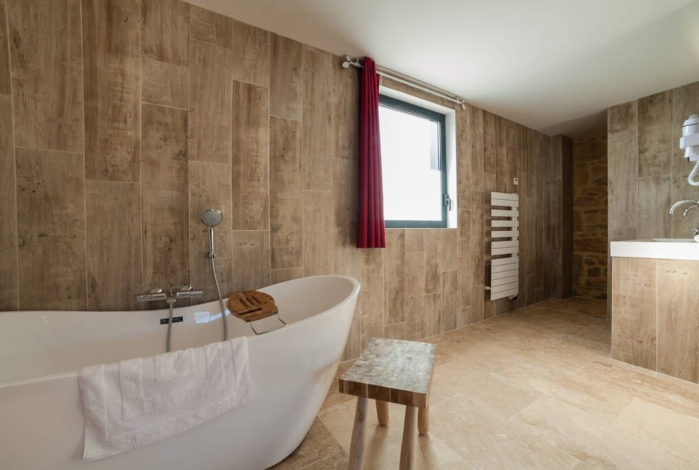 A modern bathroom with a freestanding bathtub, a small wooden stool, and a window with a red curtain.
